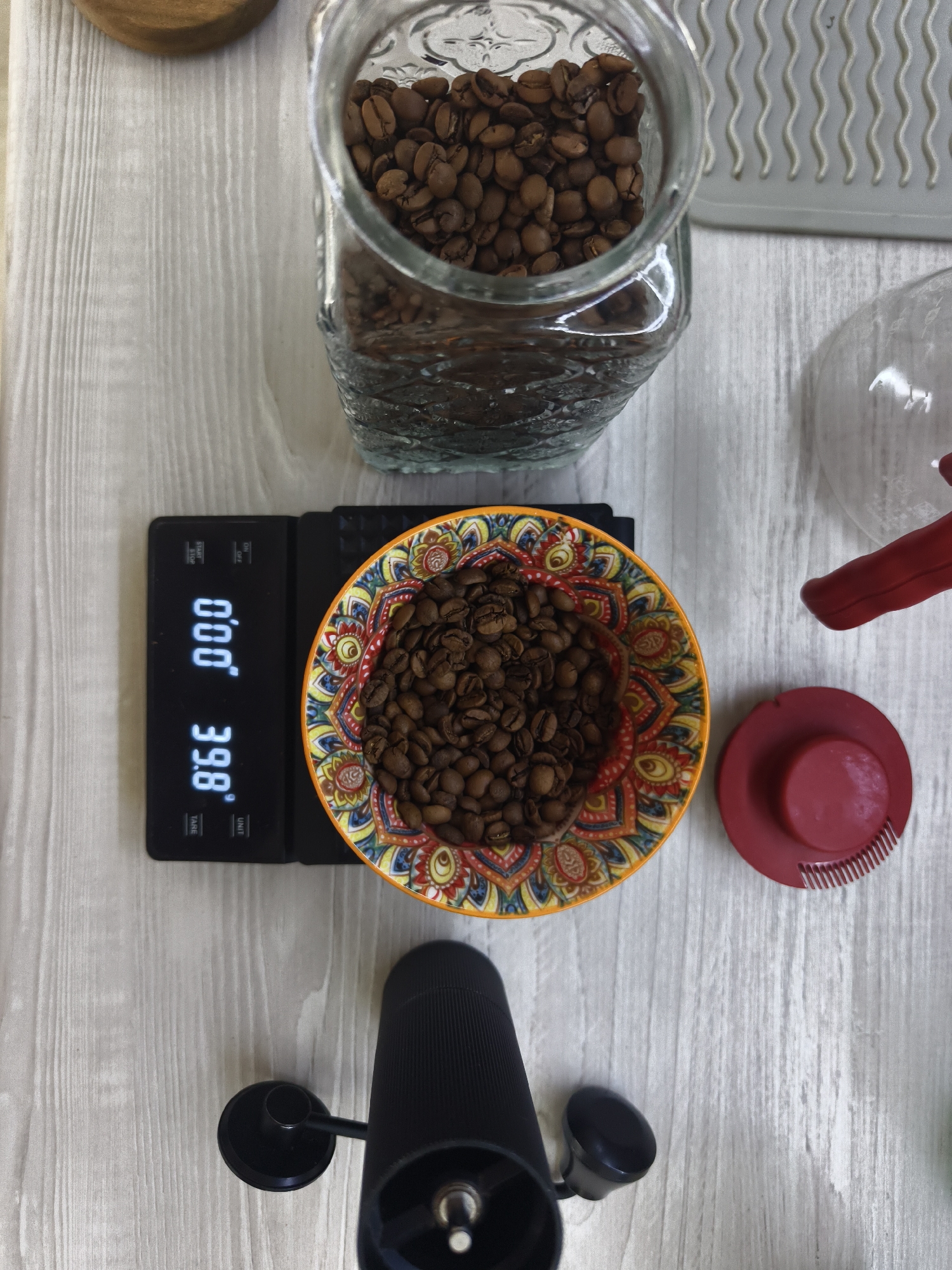 A top-down shot of a coffee preparation setup on a light gray wooden surface. The scene includes a glass jar filled with coffee beans, a black digital scale displaying "0.00", a colorful bowl containing coffee beans on the scale, a black hand grinder, and a pour-over coffee maker with a red lid nearby. The photo might have been taken indoors. The location in the city of Tyumen, Russia cannot be determined from the image.