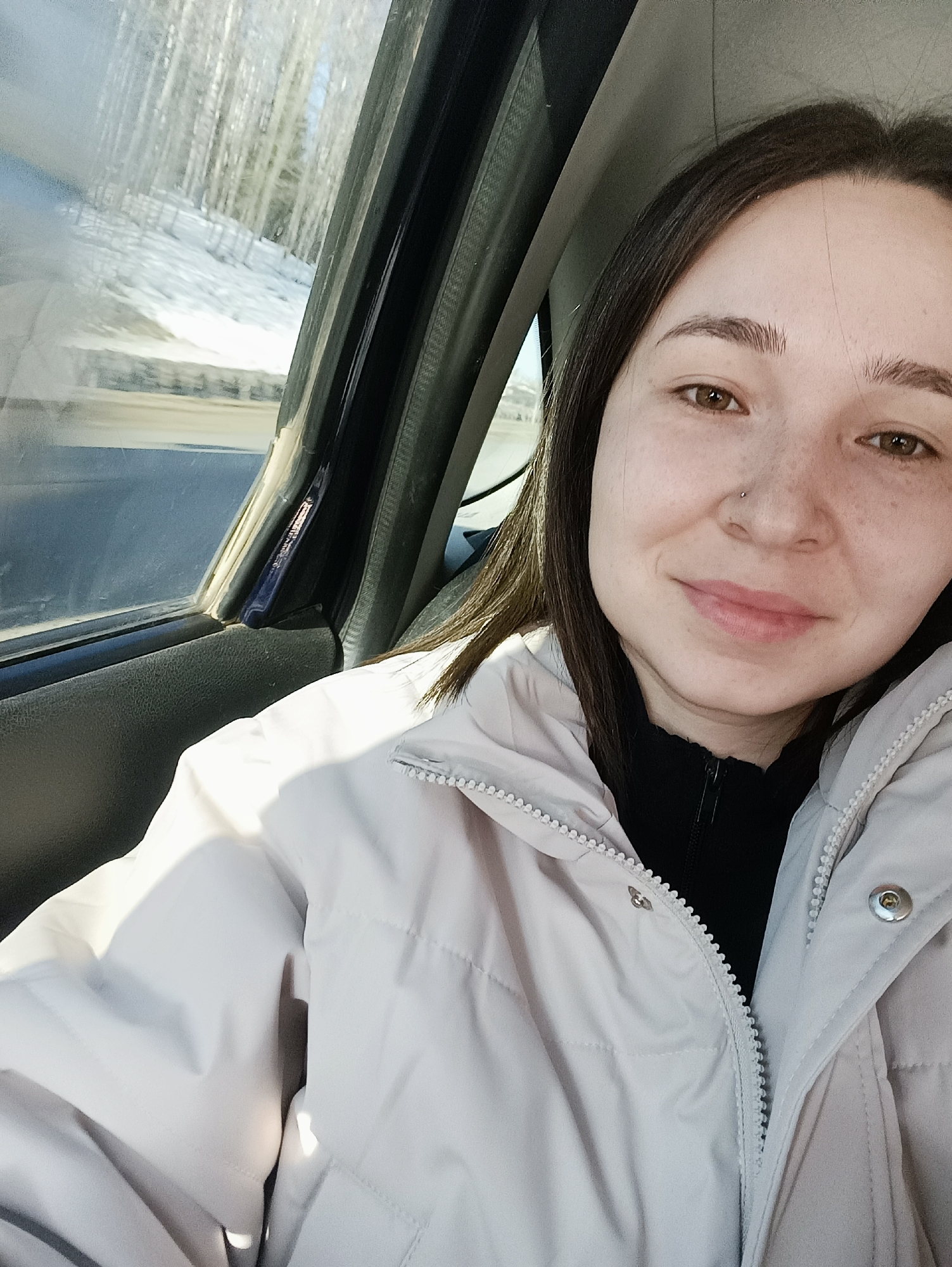 A selfie-style photograph depicts a single female subject within the passenger compartment of a vehicle, situated in Karashur, Russia. The woman, appearing to be a young adult, has dark brown hair, brown eyes, and visible freckles across her nose and cheeks. A small, spherical silver stud is present in her left nostril. Her lips are slightly parted in a faint smile. She is dressed in a light grey or beige puffer jacket, partially unzipped, revealing a black collared garment underneath. The jacket features a white zipper and snap buttons. To the left of the subject, through the vehicle's side window, a winter scene is visible. This includes a snow-covered embankment leading up to a dense stand of slender, leafless trees with light-colored bark. A blurred road surface and a metal guardrail are discernible at the base of the snowy incline. The exterior environment appears brightly illuminated by sunlight.