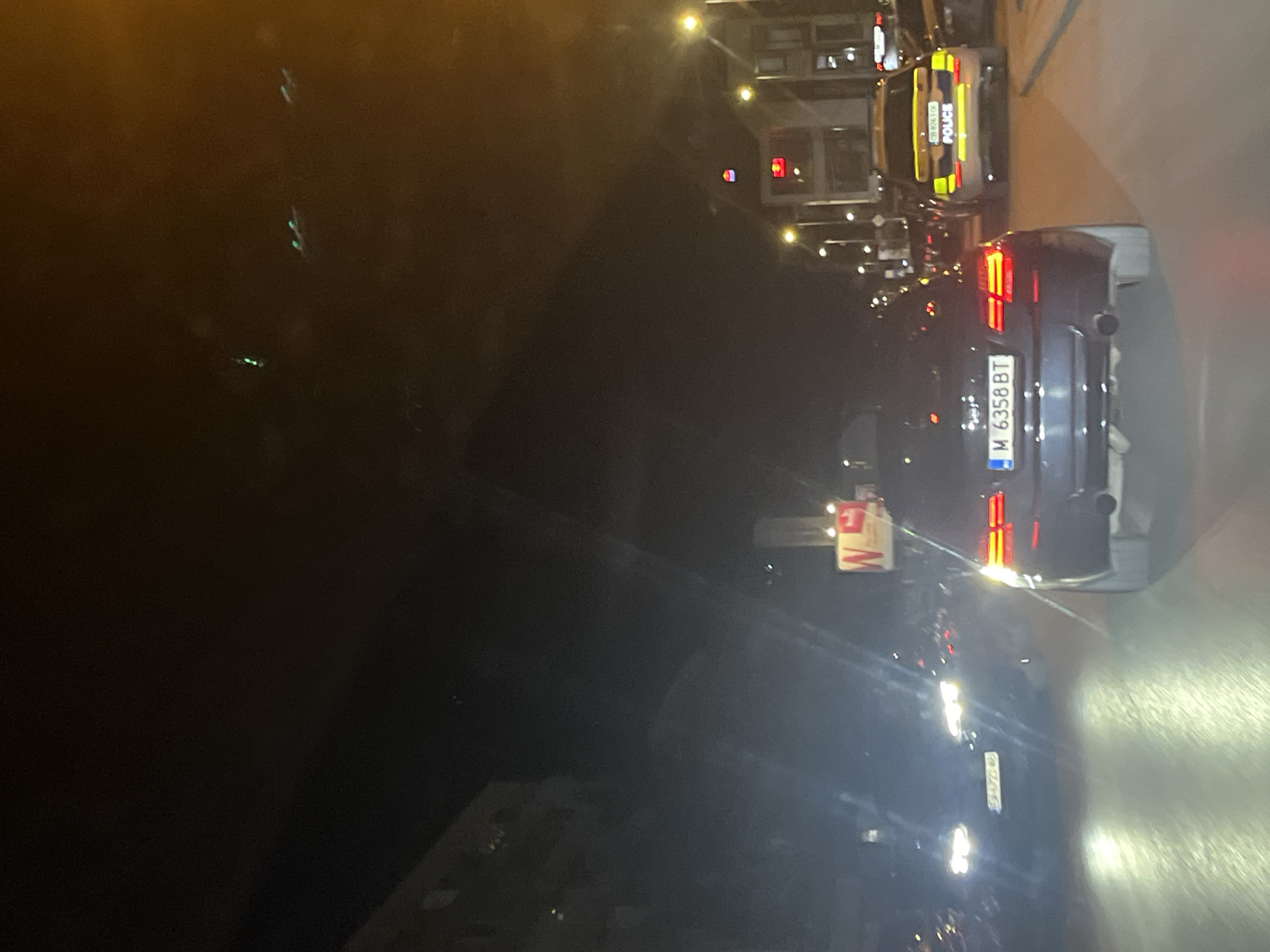The image shows a street scene at night in Sofia, Bulgaria. A black car with license plate "M 6358 BT" is visible in the foreground, its taillights illuminated. Behind it, a police van with "POLICE" written on the back is visible. Streetlights illuminate the scene. A traffic light is also visible. The environment appears to be urban.