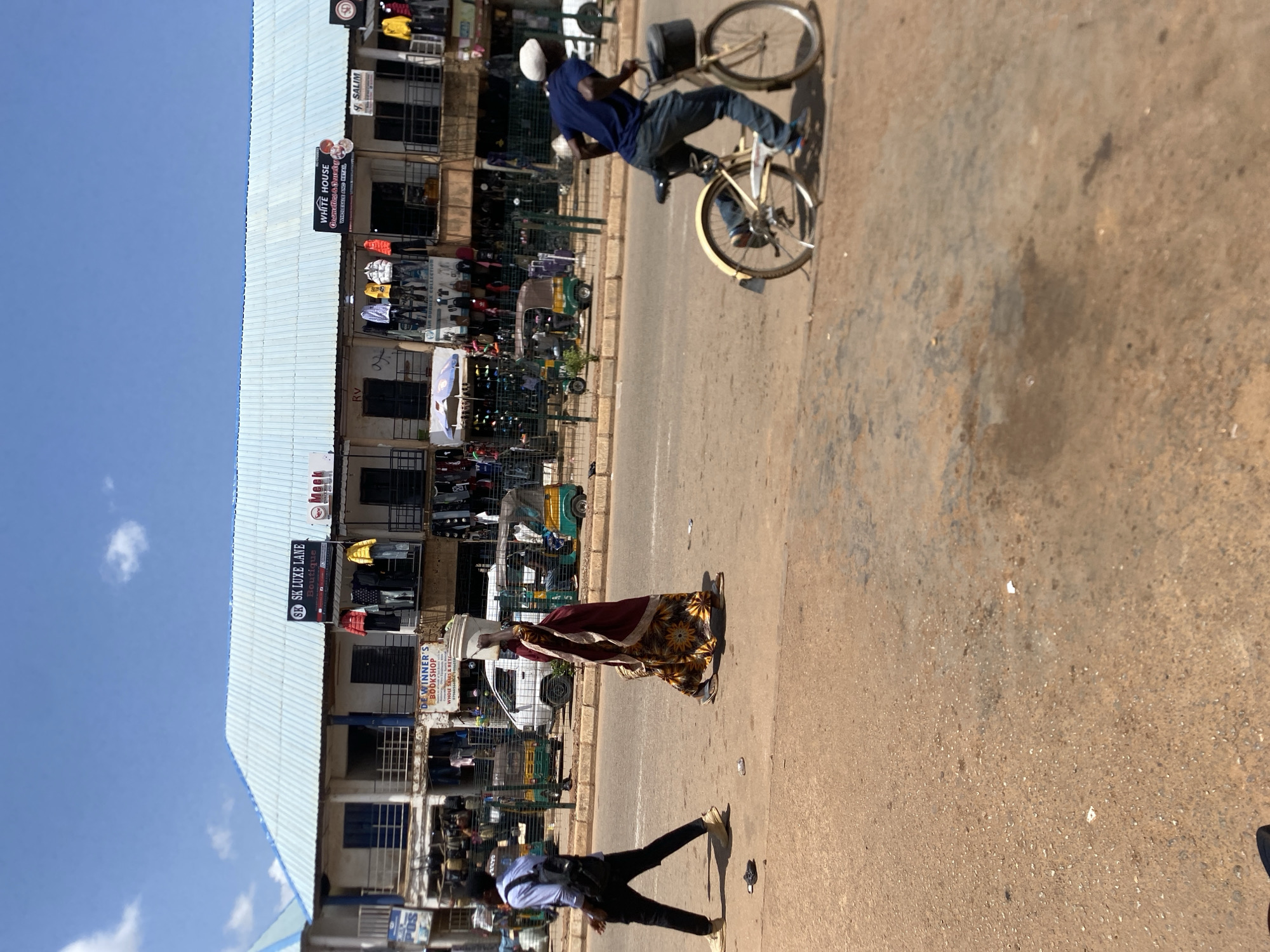 This street scene captures a vibrant day in Bukuru, Nigeria, under a clear blue sky with sparse white clouds. Bright sunlight casts distinct shadows, indicating mid-day. The image shows a paved road and a dirt shoulder, flanked by a multi-story building housing various shops, identifiable by its light blue corrugated metal roof.
On the road, a man wearing a blue t-shirt, jeans, and a white cap is actively riding a bicycle. Alongside the road, a woman in a colorful patterned dress (yellow, brown, and red) with a red shawl gracefully walks, balancing a white basin on her head. Further down the dirt shoulder, another person, seen from behind, walks while casting a long shadow.
The commercial building features several businesses. Visible shop signs include "WHITE HOUSE" advertising "Cosmetics & Jewellery WHOLESALE AND RETAIL", "SK SK LUXE LANE Boutique", and "DE WINNER'S BOOKSHOP" for "WHOLE SALES & RETAIL". Other signs bear names like "SALIM", "SD", "RV", and "NH". Clothing racks laden with merchandise are displayed outside many of the shops, and small vehicles, possibly tuk-tuks, are visible in the background, suggesting a busy commercial district. The overall atmosphere depicts everyday life and commerce.