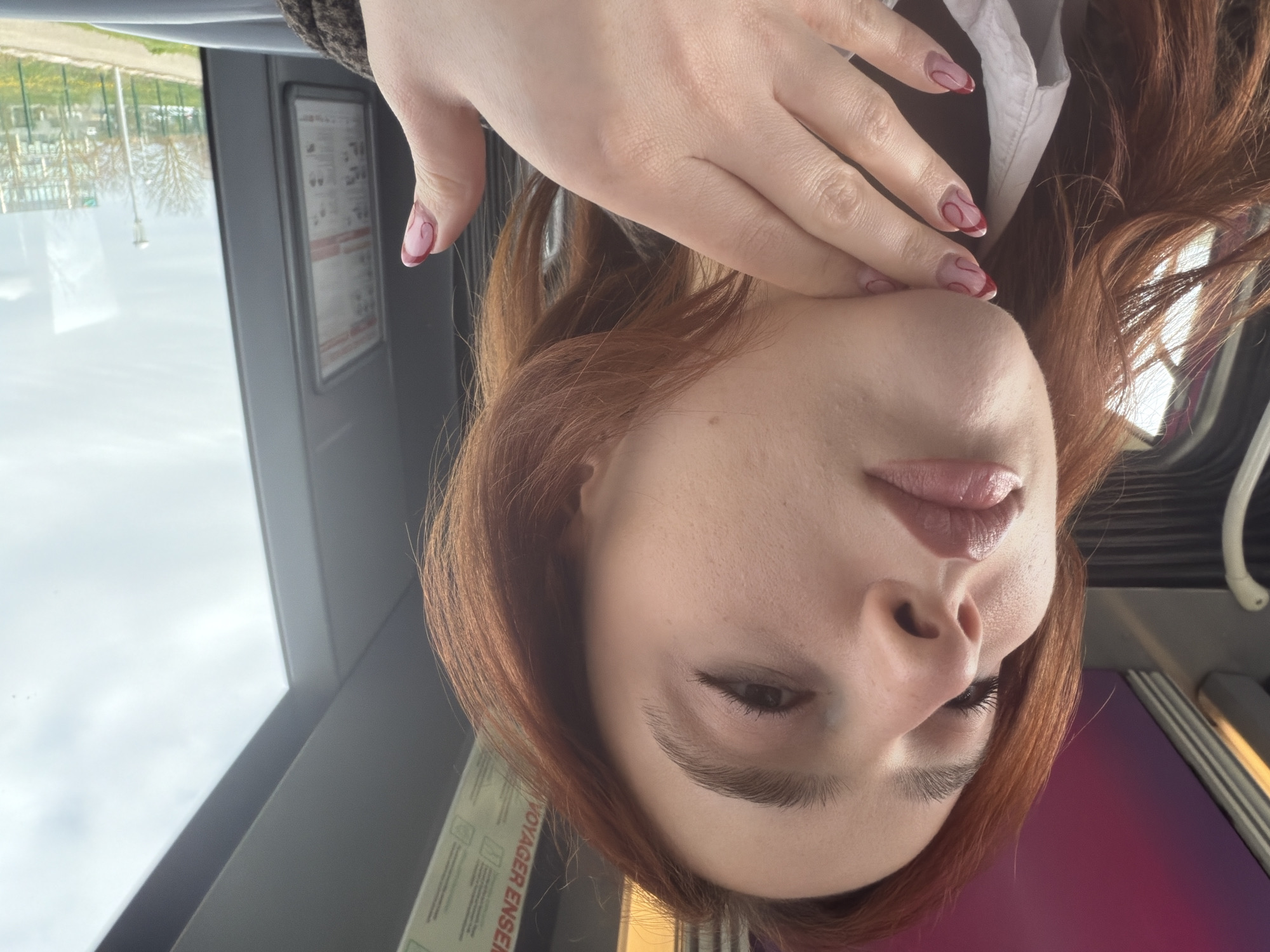 A close-up portrait shows a female subject with reddish-brown hair inside a public transport vehicle in Les Trois-Domaines, France. The subject's face is prominent, viewed from a low angle, with lips slightly parted and glossy pink. Mascara is visible on the upper eyelashes. Her left hand is positioned with fingers touching the chin/neck area; the fingernails feature clear polish with distinct red, wavy, abstract patterns. She is wearing a white collared shirt beneath a dark, textured outer garment. The vehicle's interior includes a white curved handle on the left, grey paneling, and an overhead sign partially displaying "VOYAGER ENSEMBLE." Through the window on the right, an overcast sky is visible above an outdoor scene consisting of a roadside path, a green wire fence, several leafless trees, a lamppost, and a distant multi-story building. Small patches of yellow flowers are present on the ground beside the path.