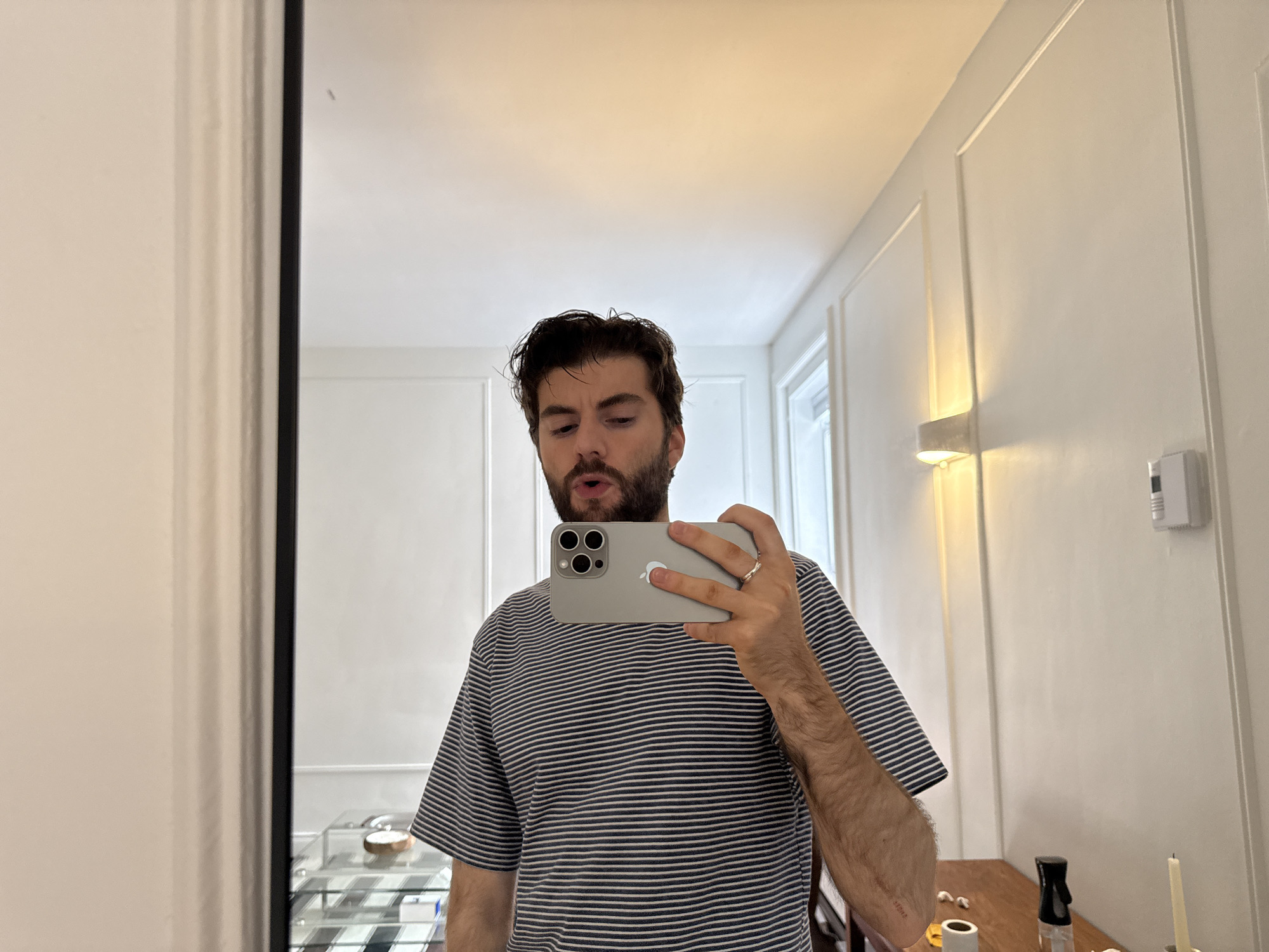 A man is taking a selfie in front of a mirror. The man has dark hair and a beard and is wearing a striped t-shirt. He is holding a gray iPhone in his right hand. The mirror reflects a room with white walls and decorative molding. There is a light fixture on the wall to the right of the man. A wooden table with various objects on it is also visible in the reflection. The location could be a residential apartment. There is no explicit information in the image to verify the location in the city of Montreal, Canada.