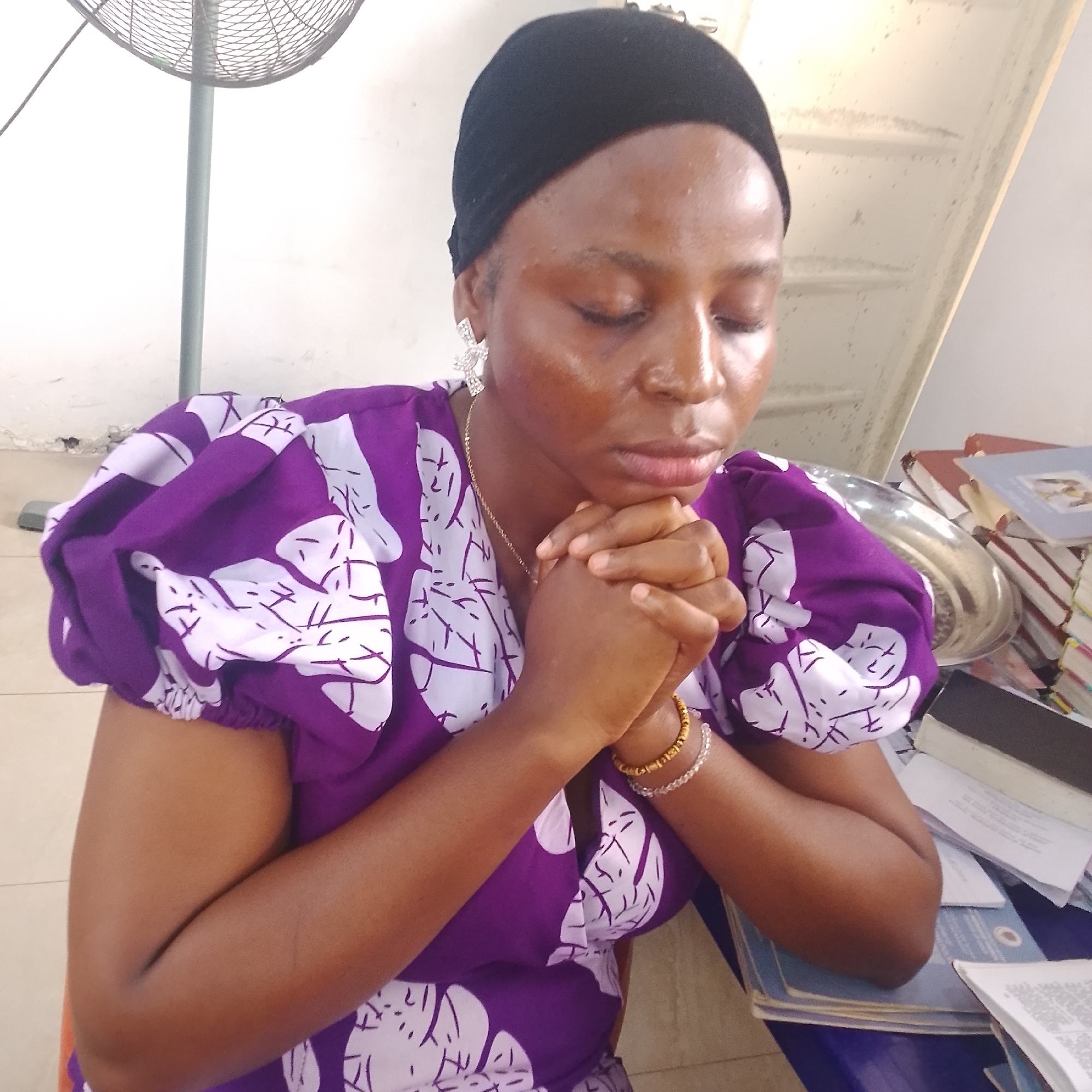 In Iranla, Nigeria, a woman is captured in a close-up shot, her hands clasped in prayer. She wears a black head covering and a vibrant purple dress adorned with a white abstract pattern. Her eyes are closed, conveying a sense of devotion. The background shows a white wall and a stacked pile of books, suggesting an indoor setting. A metal fan is partially visible to the left, and a metal tray sits on a table to the right. The lighting is soft, highlighting the woman's contemplative posture.