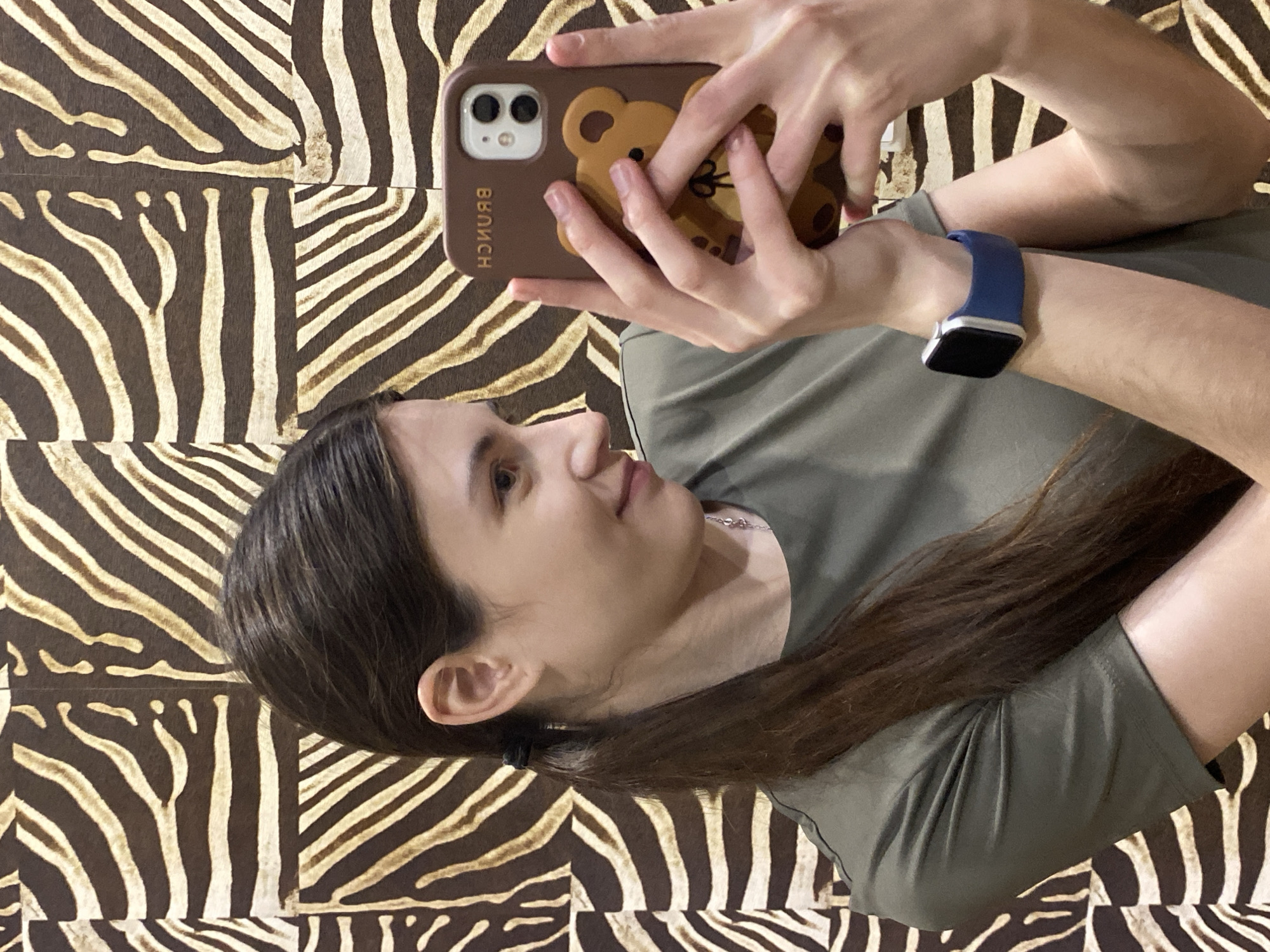 The image captures a young woman taking a selfie, holding a smartphone with both hands. Her face is shown in profile, looking slightly upwards with a calm, neutral expression. She has long, dark brown hair, which flows over her shoulder, and wears a dark olive green short-sleeved t-shirt. A silver necklace with a small pendant is visible around her neck.
On her left wrist, she wears a smartwatch featuring a black screen and a bright blue strap. The smartphone she is holding is encased in a brown protective cover adorned with a cartoon bear design. On this case, the word "BRUNCH" is distinctly visible in white capital letters, appearing mirrored due to being a reflection in the selfie. The phone's dual camera lenses are also noticeable on the upper left side of the case.
The background is characterized by a prominent zebra-print pattern, featuring irregular stripes in dark brown and light brown or beige tones. This pattern suggests an indoor setting, possibly a decorated wall, furniture, or bedding. The overall image is oriented sideways, indicating the selfie was likely taken while the subject was reclining. This scene is set in Votkinsk, Russia.