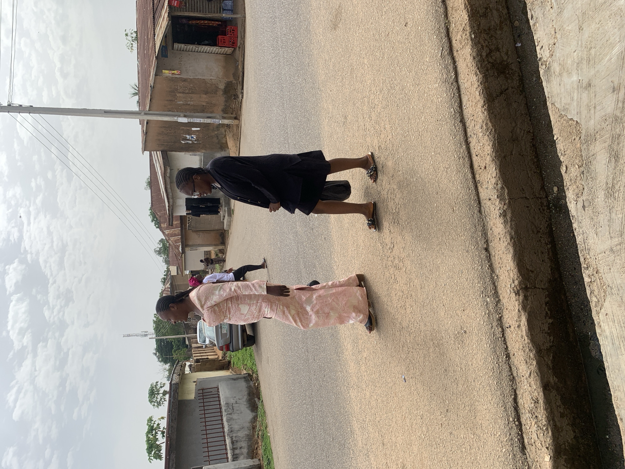 Two women walk along a paved road in Jos, Nigeria. One woman is wearing a patterned pink dress. The other woman is wearing a black outfit. They are walking in the same direction, away from the camera. In the background, there are buildings, power lines, and a parked car. The sky is cloudy. A concrete curb borders the edge of the road.