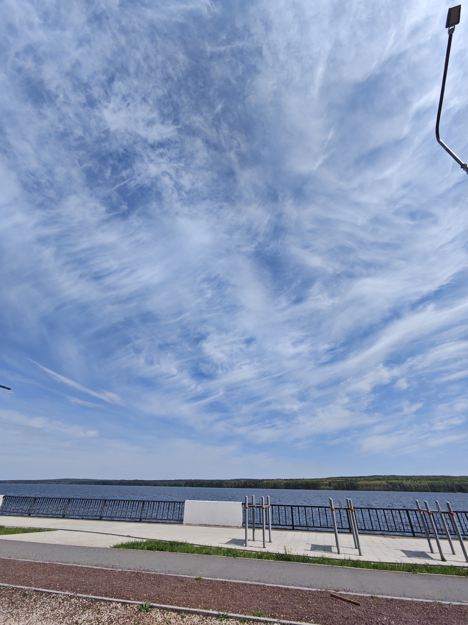 The image shows a waterfront scene in Chaikovsky, Russia. The sky is predominantly blue, filled with scattered, wispy cirrus clouds. A body of water, likely the Kama Reservoir, stretches across the midground. A line of trees is visible on the distant shore. A concrete promenade runs along the waterfront, separated from the water by a black metal fence. Exercise equipment stands along the promenade. The foreground consists of a pedestrian pathway. A light post stands on the right.