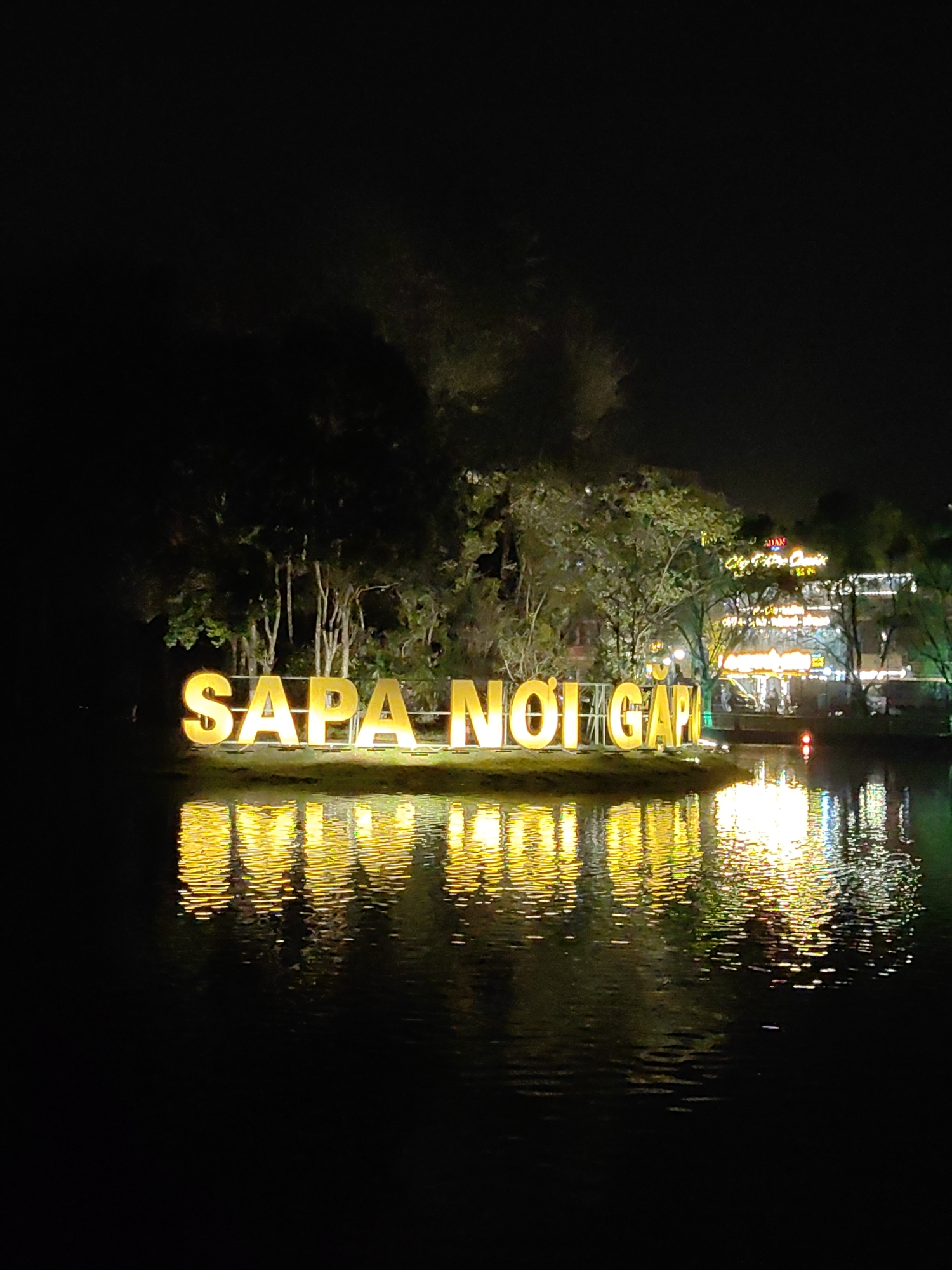 The scene depicts a nighttime view of Sa Pa Lake in Sa Pa, Vietnam. A prominent, large, yellow-illuminated sign spelling "SAPA NOI GAP" in capital block letters is situated on a small, vegetated island or landmass within the dark water. The bright illumination of the letters, along with distant lights from the shoreline, creates distinct, shimmering reflections on the rippled surface of the lake. To the left of the sign, the background consists of dark tree silhouettes. To the right, the distant shoreline features multiple-story buildings illuminated by a variety of white, yellow, and green lights, partially obscured by trees. A single red light, consistent with a buoy or marker, is visible in the water to the right of the main sign. No individuals or discernible activities are present in the image.