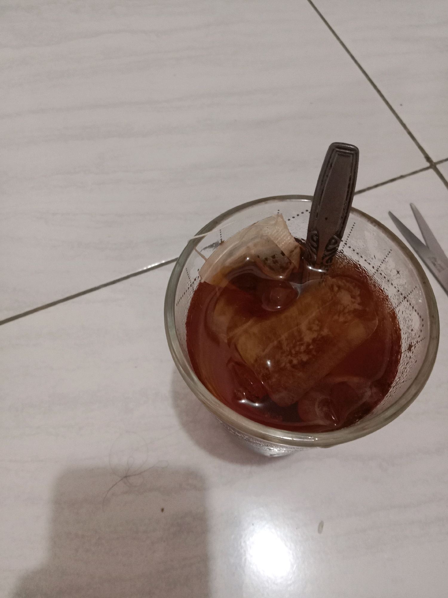 The image shows a glass of iced tea with a tea bag and a spoon. The glass is clear and filled with a dark amber liquid and ice cubes. The tea bag, made of paper, is partially submerged in the tea. A metal spoon with an ornate handle is resting in the glass. In the background, a pair of scissors is visible. The scene appears to be set on a tiled floor, likely indoors. A few strands of hair are present on the floor near the glass. The image is taken from a top-down perspective. There is no identifiable location in the city of Brangsong, Indonesia.