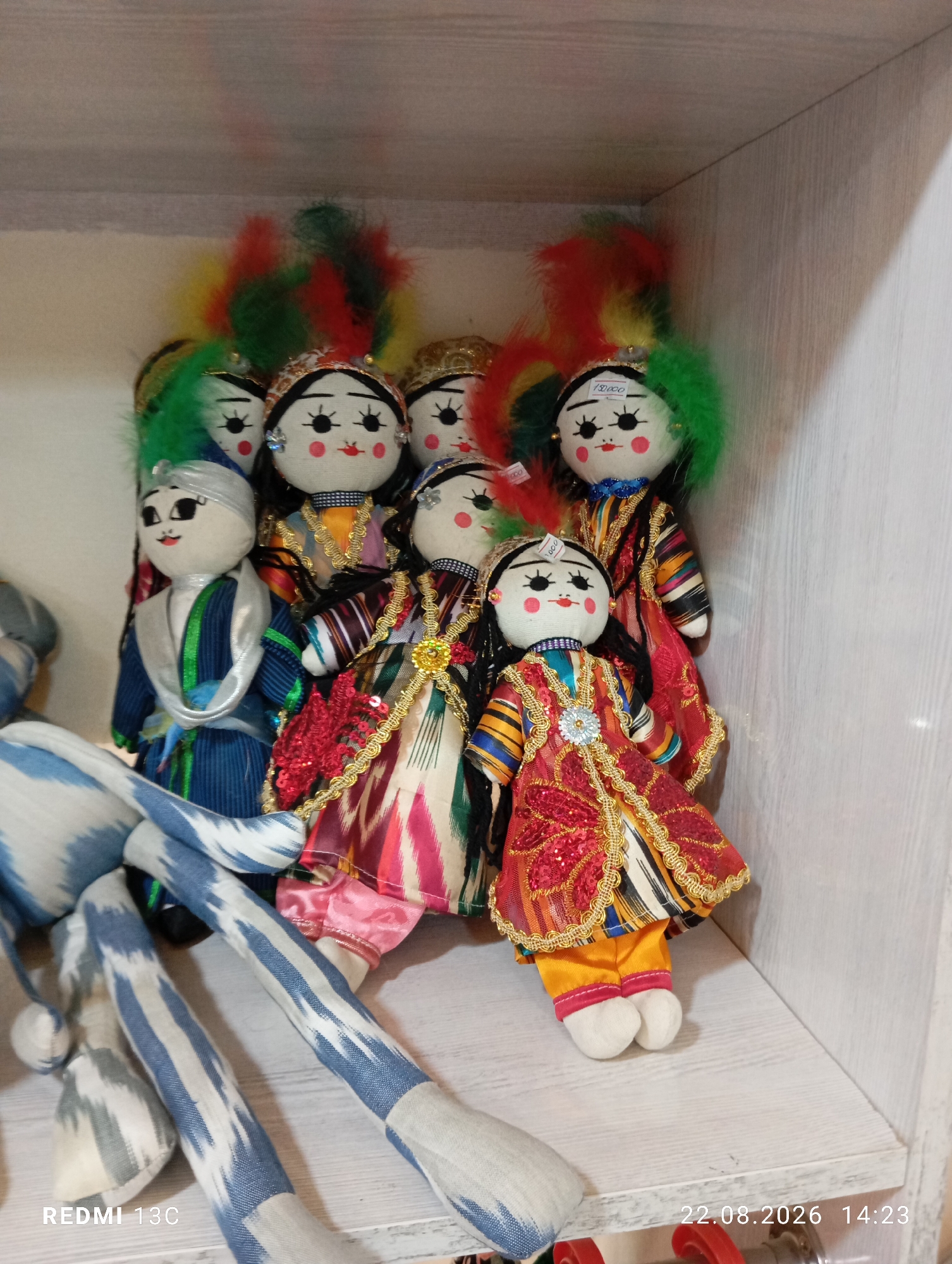 The image depicts a collection of handmade textile dolls displayed on a white wooden shelf, presumably in a shop or market setting in Amirsay, Uzbekistan. The dolls are dressed in traditional colorful clothing with intricate patterns and gold trim. Several dolls have bright feather headdresses in red, yellow, and green. One doll, positioned in the front right, wears a vibrant red dress with gold embroidery and a yellow outfit underneath, along with white socks and red trim on its pants. Another doll on the left wears a blue and green robe with a silver sash and head covering. In the foreground, partially visible, are the legs of a large plush toy or doll made of fabric with a blue and white ikat-like pattern. A price tag, reading "150000", is visible on the headdress of one of the dolls. The lighting suggests an indoor environment. The timestamp "22.08.2026 14:23" is present in the lower right corner, and "REDMI 13C" is in the lower left, indicating the device used for capture.