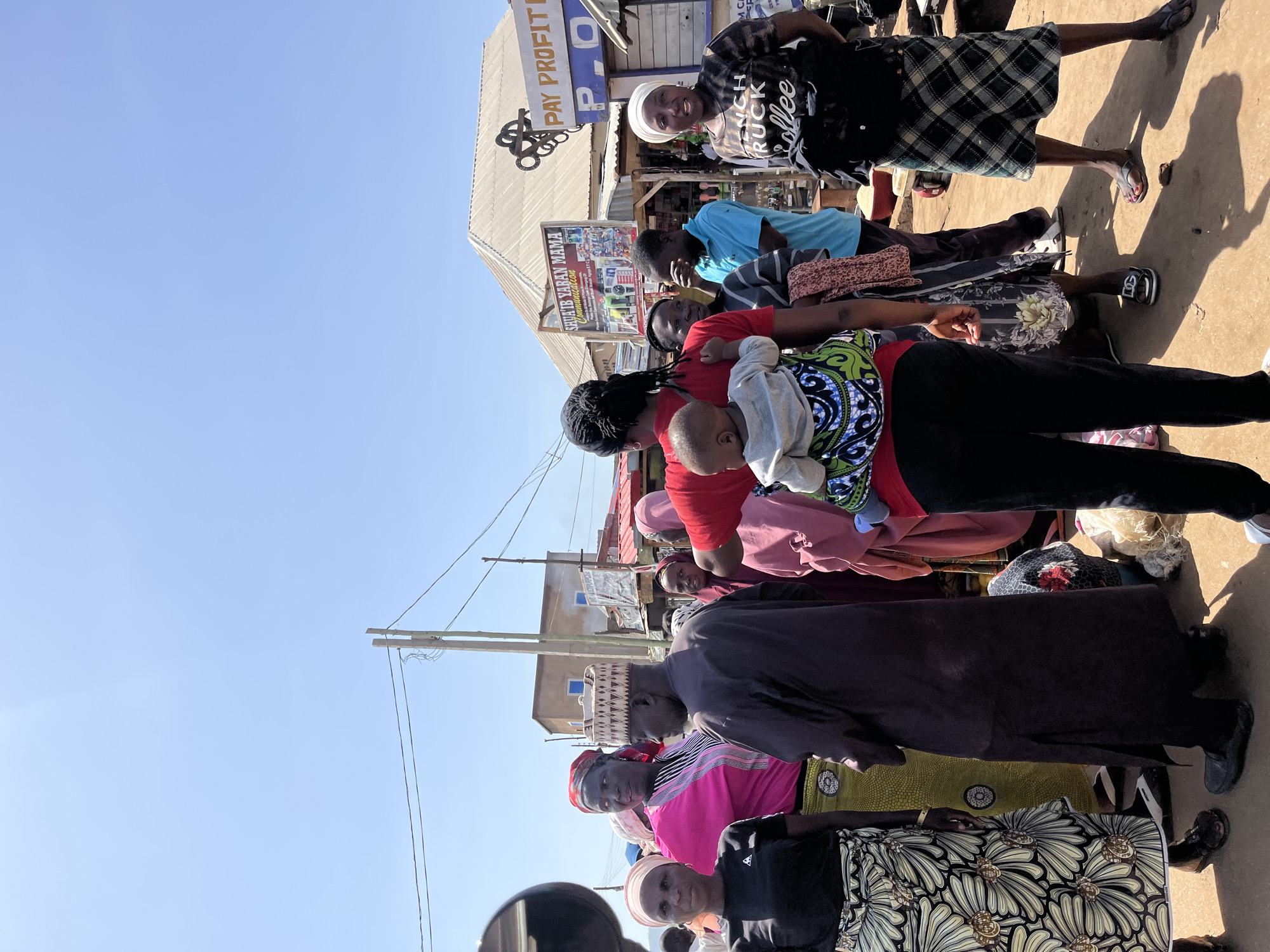 This image depicts a bustling street scene in Rafin Par, Nigeria, under a bright, clear sky. The time of day appears to be mid-morning or late afternoon, as strong sunlight casts distinct shadows on the sandy ground.
Numerous people are gathered, primarily women and children, engaged in what appears to be daily activity or a communal gathering. In the foreground, a woman wearing a red top and black trousers carries a baby securely on her back in a patterned fabric carrier, featuring blue and green designs. Nearby, another woman is dressed in a black and white patterned skirt and a dark top. Further back, a woman with a white headwrap and a plaid skirt is smiling towards the camera. Her black t-shirt displays white text, which reads "CRUSH TRUCK coffee." Among the crowd, a man in a blue shirt and a traditional patterned cap is also visible. The individuals wear a variety of clothing, ranging from traditional patterned fabrics and head coverings to more contemporary attire.
The background features several buildings, likely shops or market stalls, characterized by their corrugated metal roofs. Visible text on signs includes "PAY PROFIT" and "P O" on one sign, and "SHU'AIB YARA NAMA C..." (likely part of a longer business name such as "Communications") on another. A wooden utility pole with wires runs through the upper right portion of the frame, reinforcing the urban street setting. The scene conveys an impression of everyday life and interaction in a lively outdoor environment.