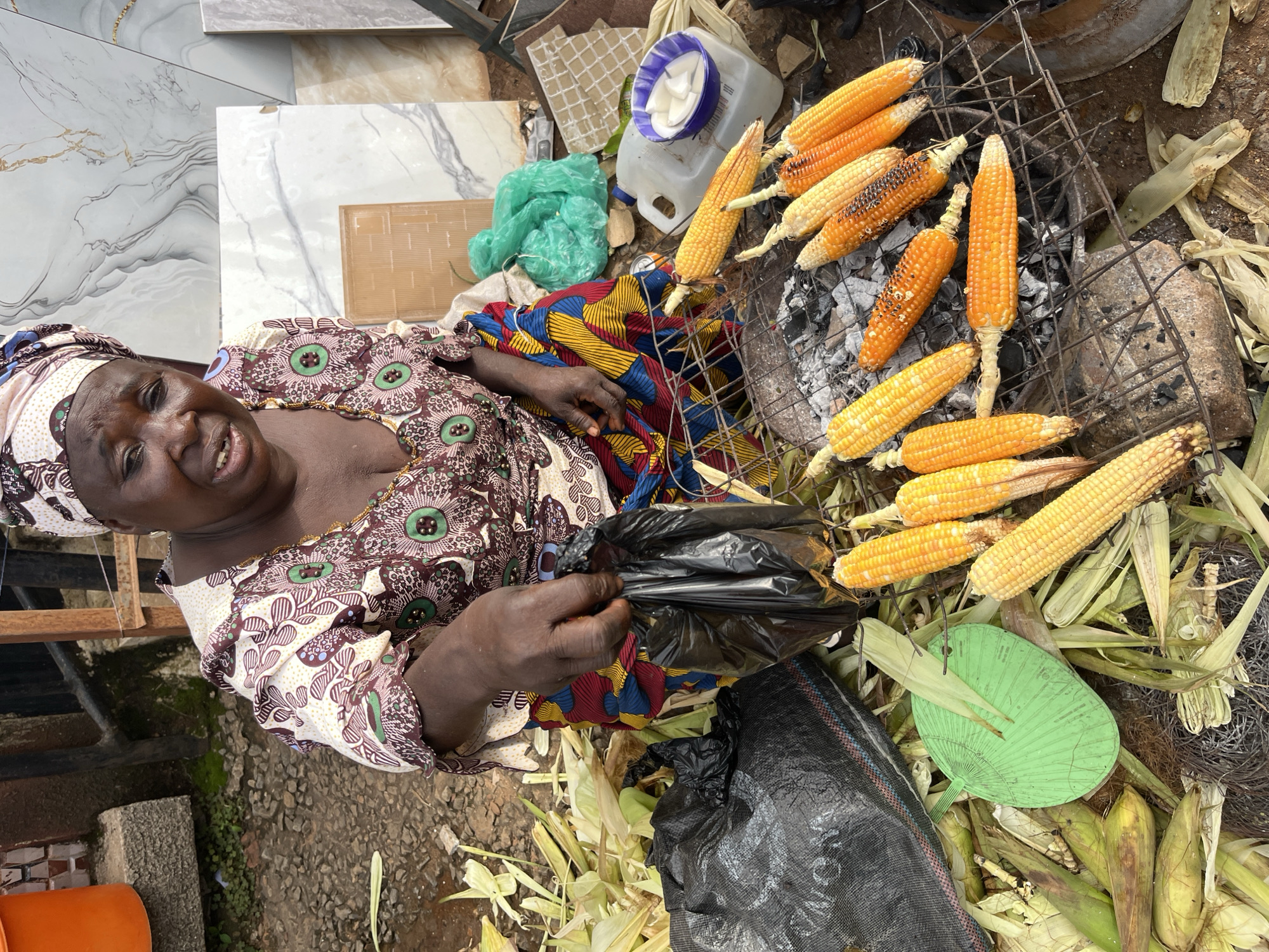 A woman wearing a colorful headwrap and a patterned dress is shown in what appears to be an outdoor market stall in Gura Top, Nigeria. She is looking towards the camera with a slight smile. In front of her, corn is being grilled on a metal rack over charcoal. Several ears of corn, some partially charred, are arranged on the grill. Piles of husks and more ears of corn are visible around the grilling area, along with a green fan, a plastic container with white contents, and some green plastic bags. The ground is a mix of dirt and scattered debris. The scene suggests a food preparation or vending activity, likely taking place during the day. The overall impression is one of a busy, informal market setting.