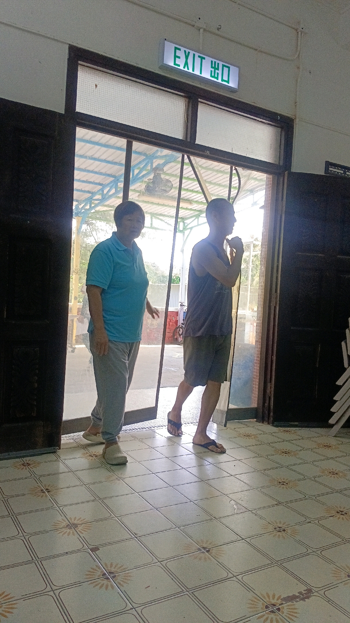 An eye-level shot shows two individuals, one woman and one man, standing and walking through a doorway. The woman, on the left, is wearing a blue polo shirt and grey pants. The man, on the right, is wearing a dark tank top and grey shorts. Both are barefoot. Above the doorway, a green illuminated "EXIT" sign with Chinese characters is visible. The environment appears to be an indoor space with tiled flooring and wooden doors. The scene suggests a casual setting, possibly a community center or a public building in Hong Kong, China, given the bilingual signage.