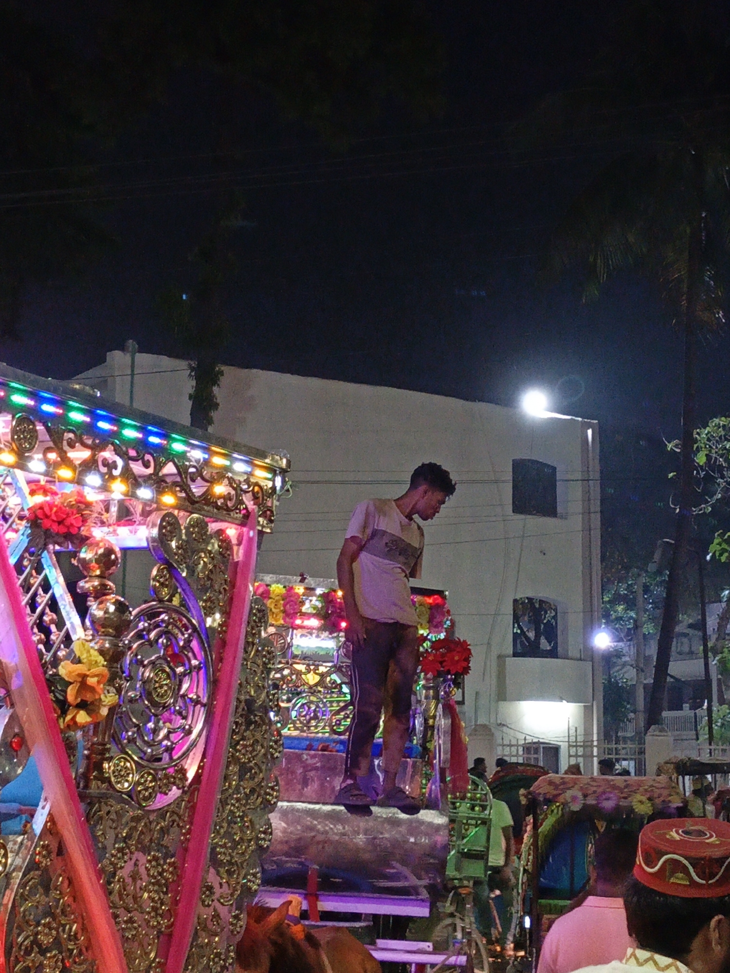 The image depicts a nighttime scene in Dhaka, Bangladesh. A man stands on a decorated horse-drawn carriage. The carriage is adorned with ornate designs, bright lights, and colorful flowers. Other people are visible in the crowd, with at least one wearing a red kufi. The scene is illuminated by streetlights, with a white building in the background.