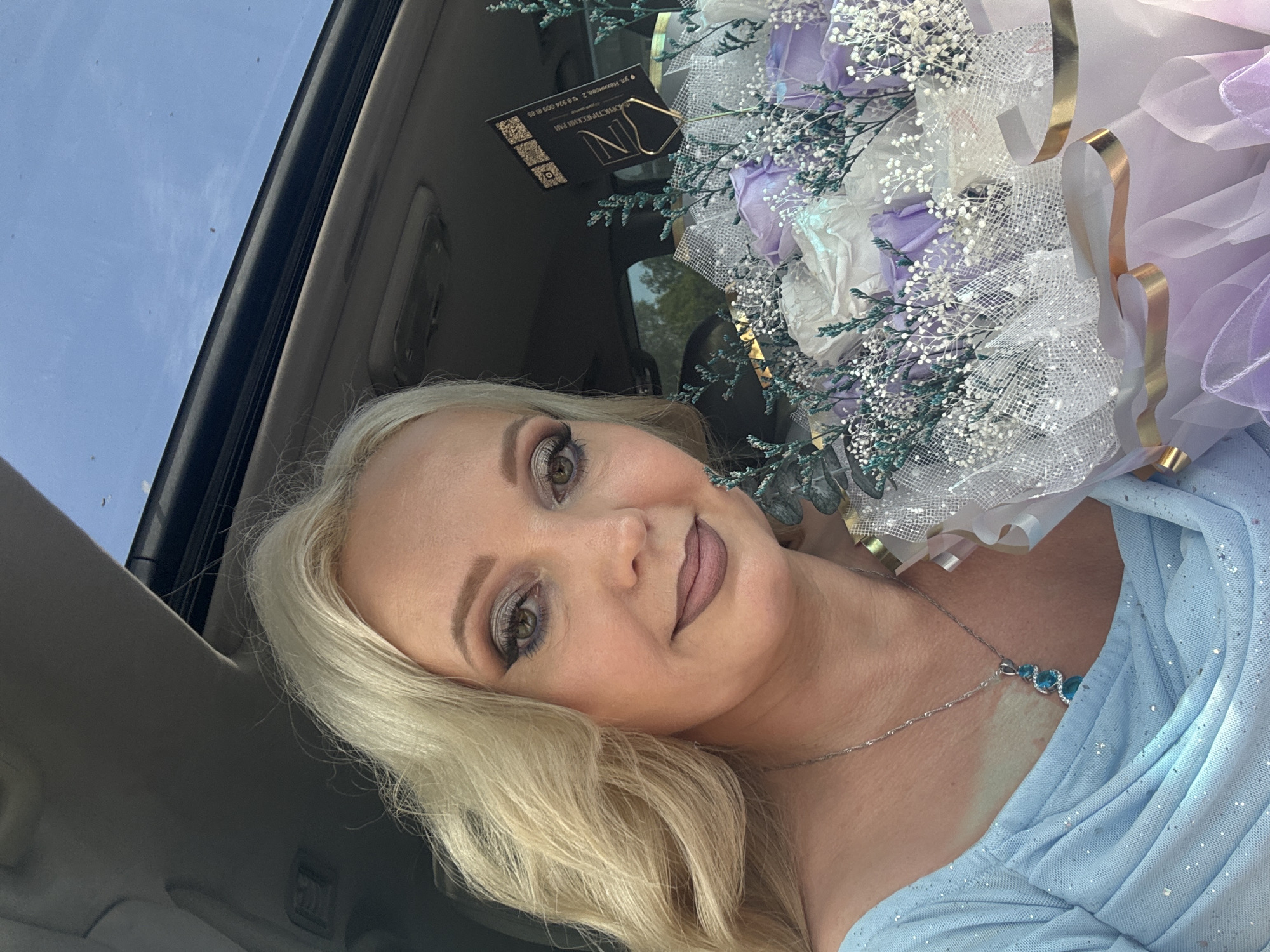 A blonde woman with makeup is sitting in a car. She is holding a bouquet of purple and white roses, and has a blue pendant necklace on. She is wearing a light blue dress. The car has a sunroof open, and the sky is visible through it. A business card is attached to the bouquet. The scene appears to be in Komsomolsk-on-Amur, Russia, based on the text on the business card which includes a Russian address.