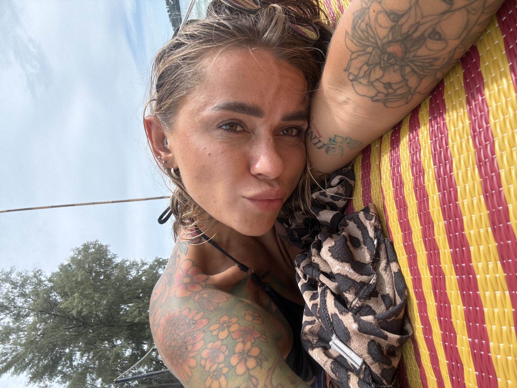 A woman with extensive tattoos on her arms and chest lies on a red and yellow striped mat outdoors, likely in Choeng Thale, Thailand. She is resting her head on her arm, and her gaze is directed towards the viewer. A piece of leopard-print fabric is folded beside her. In the background, a large tree with green foliage and a partly cloudy sky are visible, suggesting an outdoor, possibly beach or coastal, setting.