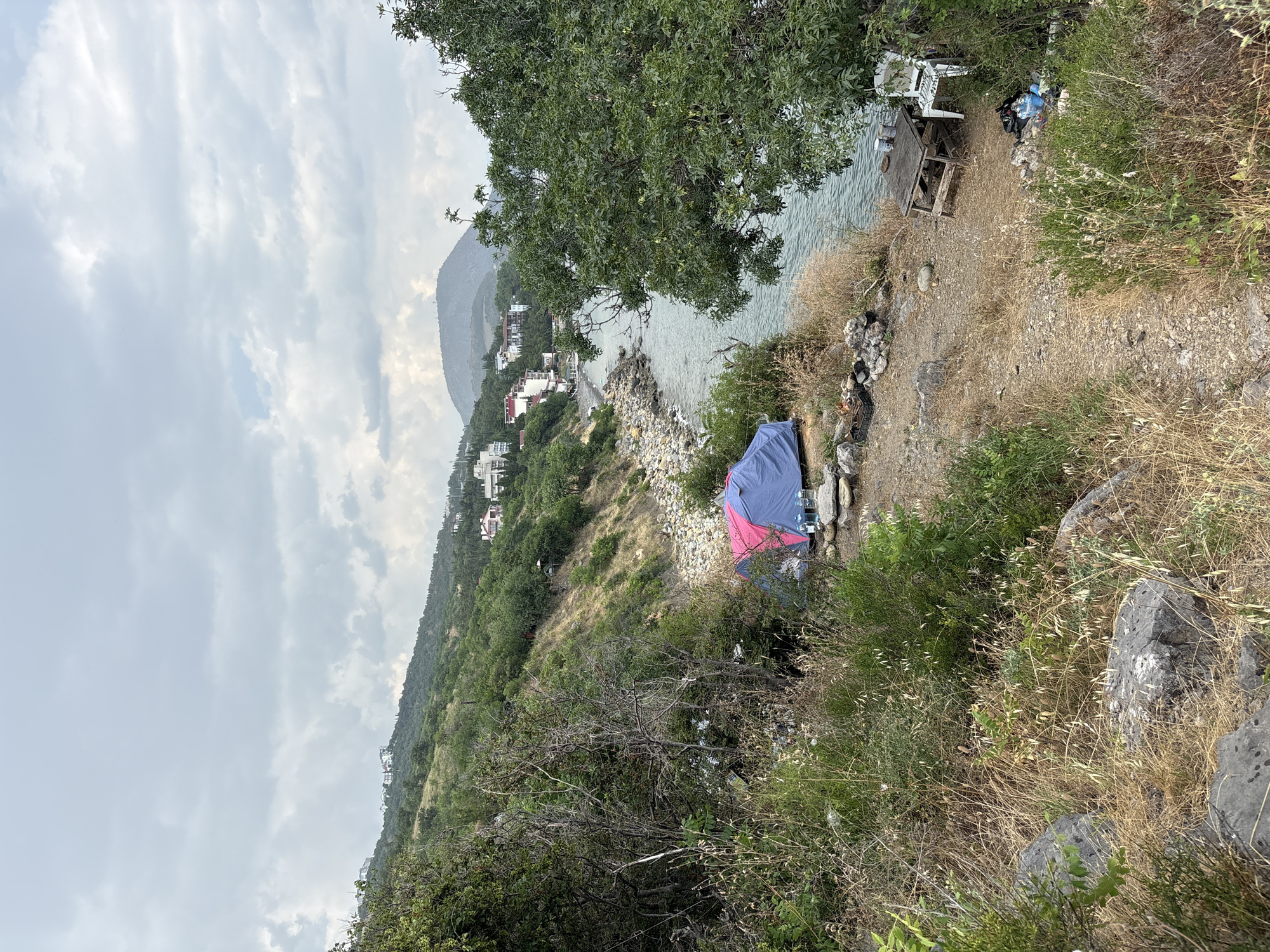 A view of the coastal area in Bondarenkovo, Ukraine, shows a campsite near the sea under a cloudy sky. A blue and pink tent is set up on a rocky, grassy area. In front of the tent, there are several bottles of water. Nearby, a rustic wooden table and a white plastic chair are placed, with trash bags and plastic containers scattered around them. The sea stretches out to the right, with a rocky shore leading up to a hillside covered in green vegetation and scattered buildings. The overall scene suggests a somewhat untidy and temporary dwelling.