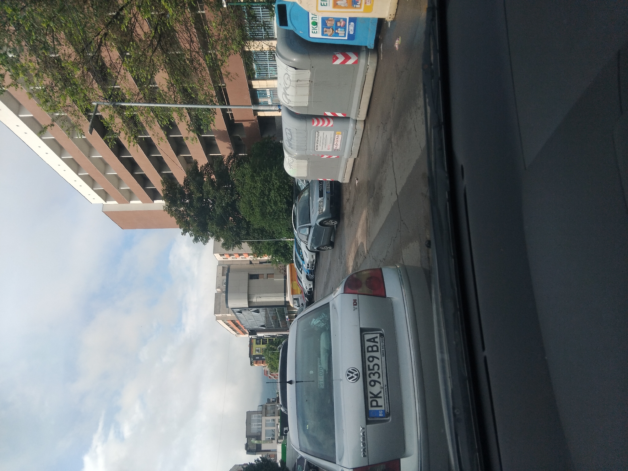 The image presents an urban street scene, likely captured from inside a vehicle, looking towards the curb and buildings. The setting appears to be a residential or mixed-use area in Sofia, Bulgaria, identifiable by the Bulgarian text and license plate details.
In the foreground, the rear of a silver Volkswagen Passat car occupies the bottom portion of the frame. Its Bulgarian license plate, "PK 9359 BA," is clearly visible, along with smaller text reading "www.cartmax.mobile.bg" and "0898 602 002." A "TDI" badge is on the right side of the trunk, and "PASSAT" is embossed on the left. A sticker on the rear window displays "ST-070-MMA."
Behind the Passat, a row of other parked cars—including grey, white, and blue models—can be seen. Adjacent to these vehicles, a series of waste containers lines the street. Two prominent grey bins feature red and white striped reflective tape and circular markings, with one bearing the Cyrillic inscription "СМЕСЕНИ БИТОВИ ОТПАДЪЦИ" (Mixed household waste). Beside them is a blue recycling bin, branded "EKOPAK," displaying universal symbols for sorting different types of recyclables like glass, paper, and plastic.
The background consists of several multi-story buildings with brown and white facades, some featuring balconies. Trees with green foliage are interspersed throughout the scene, adding natural elements to the urban landscape. Above the buildings, the sky is partly cloudy but bright, indicating a daytime setting, possibly morning or afternoon. There are no people visible, and no specific activity or event is taking place, depicting a typical quiet street view.