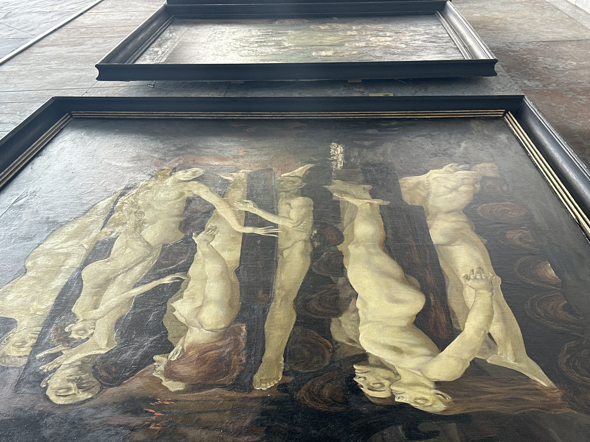 The image shows a close-up of a painting, likely part of an exhibition, in Minsk, Belarus. The painting depicts several pale, nude figures in various states of repose or distress, layered horizontally. The style is somewhat abstract and somber, with dark backgrounds and muted tones. The figures appear to be sleeping or possibly deceased, with limbs intertwined and faces turned away or down. In the foreground, two figures are prominently displayed, their bodies interlocking. To the right, another framed painting is partially visible, showing a crowd of figures rendered in a more impressionistic style. The paintings are displayed on a light-colored, textured wall.