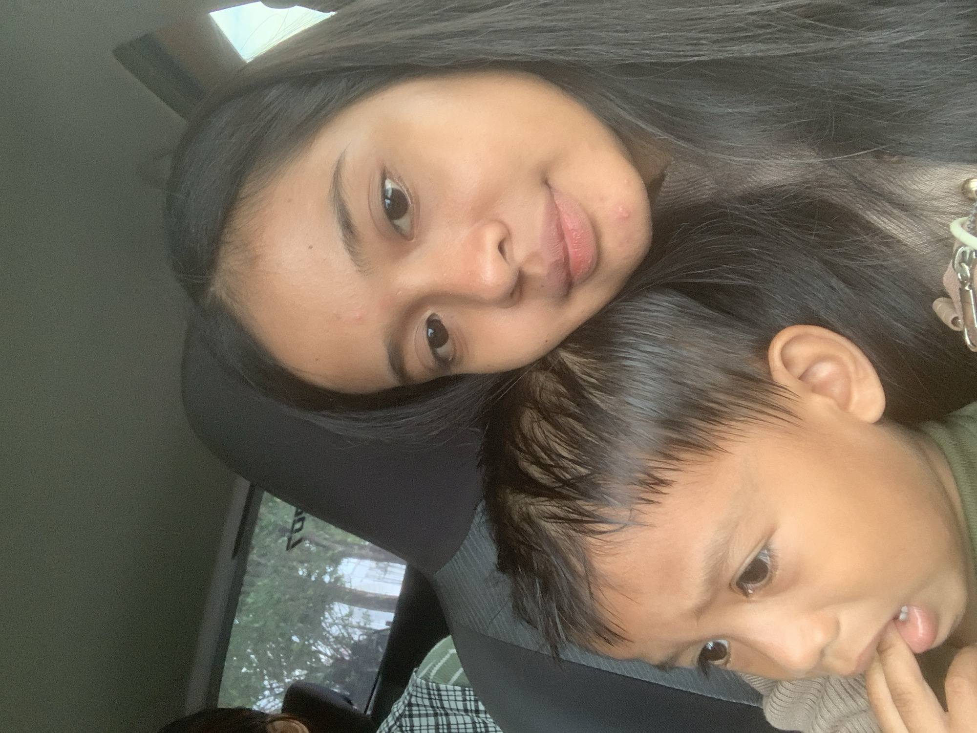 Two individuals are visible in the image: an adult female and a young child, presumably male. The female is positioned behind and slightly above the child. Her head is angled towards the camera, with her dark hair spread around her head and shoulders. Her facial expression appears neutral. The child is positioned below her, facing slightly to the right of the frame. The child has their finger in their mouth. Both appear to be inside a vehicle, the details of the interior visible in the background, including a window and part of a car seat. Given the prompt, the location is implied to be within the city of Samarinda, Indonesia.