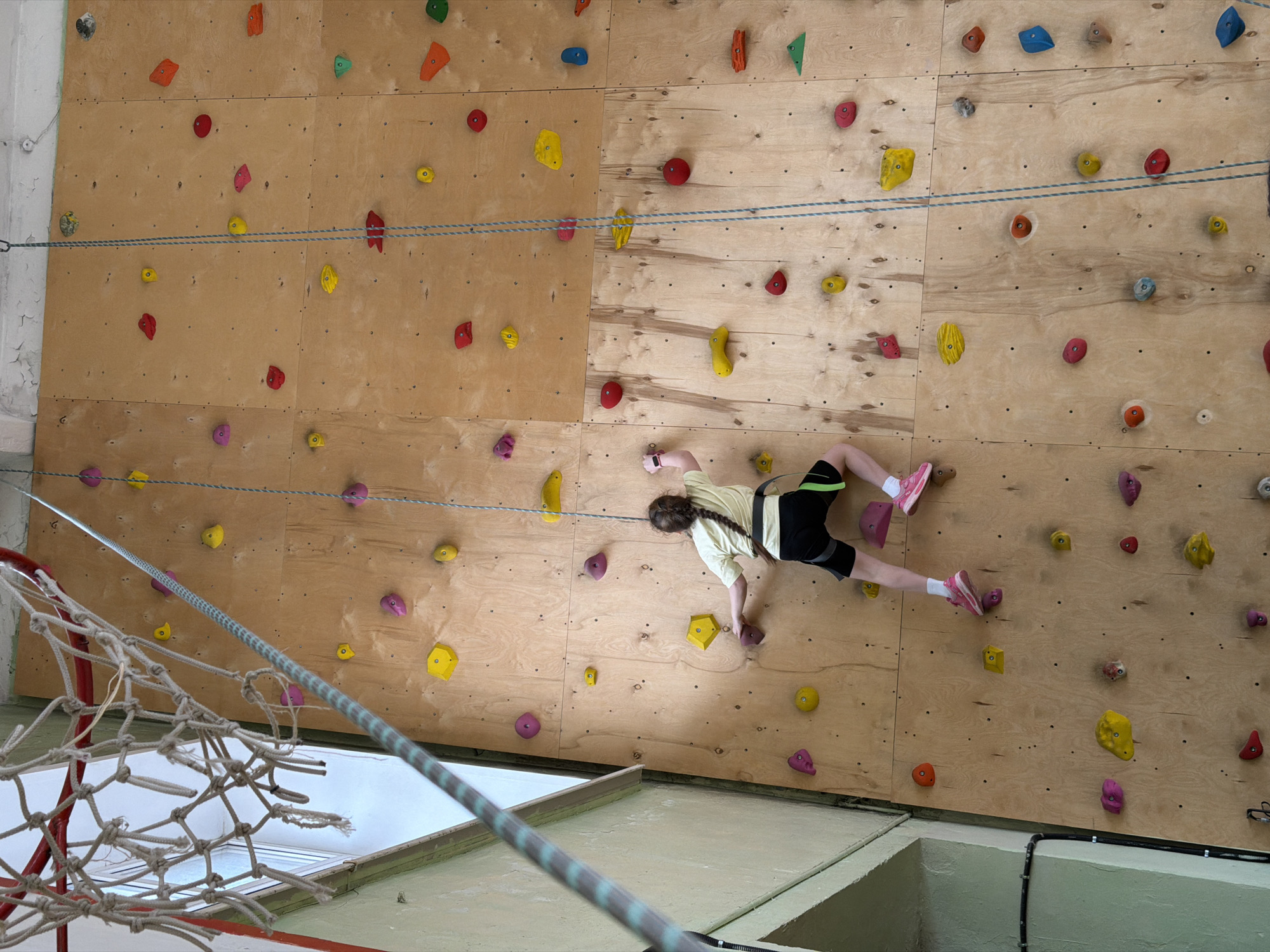 A young girl is rock climbing indoors. She is ascending a wooden climbing wall covered with various colored handholds. The girl is wearing a light yellow shirt, black shorts, and pink shoes. A safety rope is visible, extending from the top of the wall down to the bottom. A net is in the lower-left corner of the image. The climbing wall is in Izhevsk, Russia.
