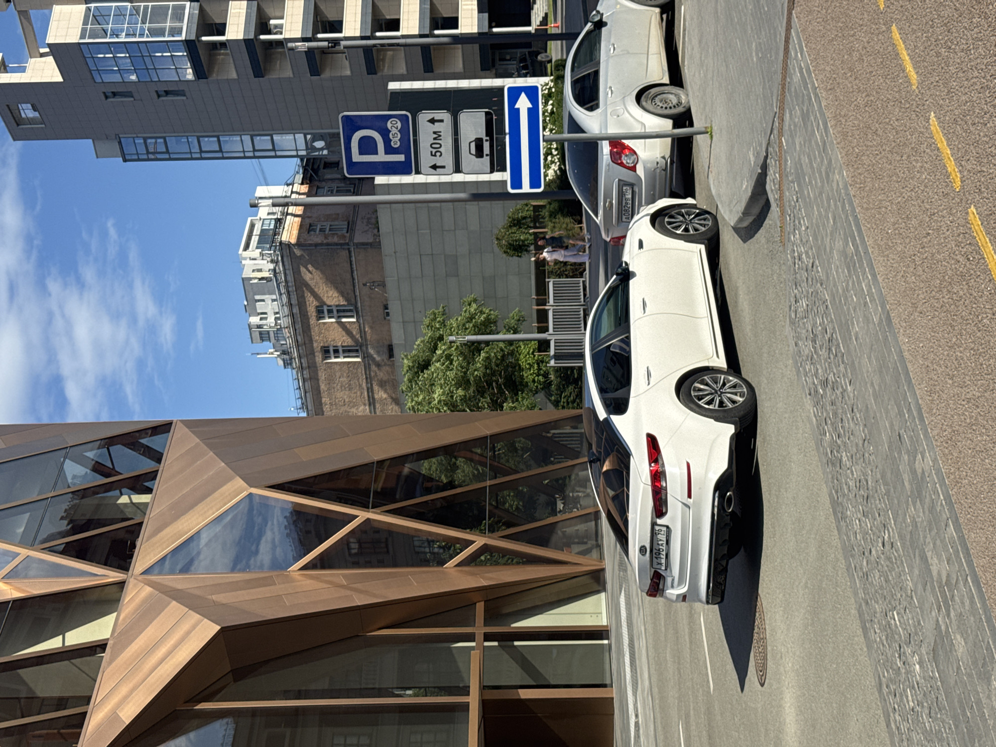 The image shows a street scene in Yekaterinburg, Russia. In the foreground, a modern building with a geometric, faceted facade made of copper-colored panels and glass is visible. To the right, two white cars are parked along the street. The car in the foreground is a white sedan, and behind it is another white car, possibly an SUV.
Several traffic signs are mounted on a pole. The signs include a blue parking sign with a "P" and "10-20" indicating parking hours, followed by a sign showing an upward arrow with "50m," and a sign with a truck symbol. The furthest sign is a blue rectangular sign with a white upward arrow.
In the background, there are several buildings. One is a tall, modern building with many windows, and next to it is an older, brick building. Trees are also visible, with lush green foliage. The sky is blue with some white clouds.
Two people are standing near the parked cars, seemingly conversing. The lighting suggests it is daytime, likely mid-morning or afternoon, with bright sunlight casting shadows. The license plate on the foreground car is visible and reads "X 196 AY 96". The overall impression is a bright, sunny day in an urban environment.