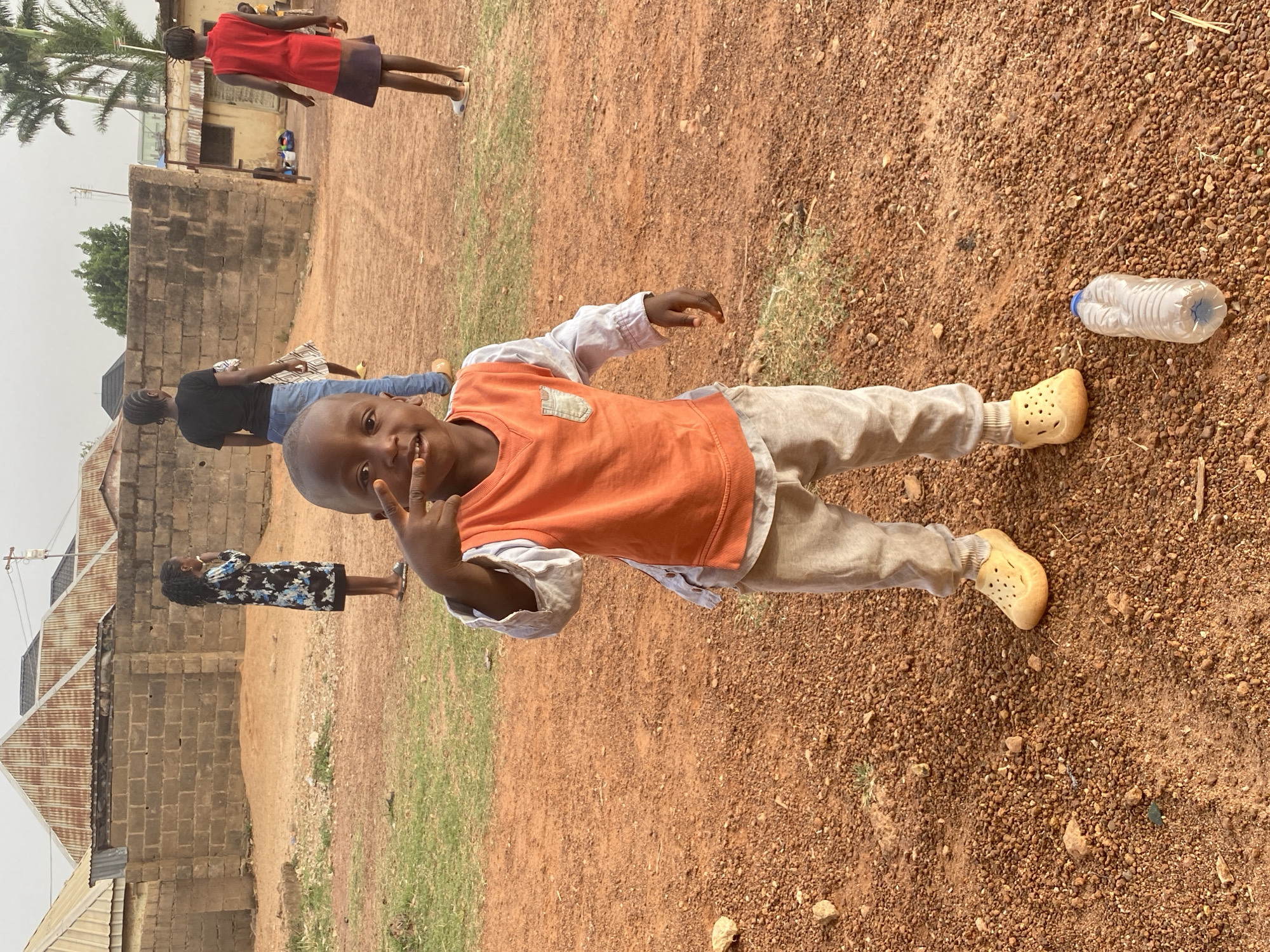 The image, taken in Bukuru, Nigeria, features a cheerful young child as the primary subject, standing in an outdoor setting and posing for the camera. The child, a boy with dark skin, is in the foreground, smiling broadly and making a peace sign with his right hand. He is dressed in a bright orange sleeveless top worn over a light-colored long-sleeved shirt, paired with light-colored long pants and light-colored slip-on shoes resembling Crocs.
In the midground and background, several other people are visible. To the left, a person in a black top and blue jeans is partially seen, and further back, a woman in a black and white patterned dress stands, looking away from the camera. To the right, another individual in a red dress with a dark lower half walks away, with a glimpse of a person in a yellow top behind them.
The scene takes place on reddish-brown, unpaved ground covered with small stones and sparse patches of dry grass. In the background, simple buildings constructed from concrete blocks and topped with corrugated iron roofs indicate a village or rural environment. A tall palm tree and other green vegetation are also present. A clear plastic water bottle with a blue cap lies on the ground near the foreground child. The bright, even lighting suggests it is daytime. The prominent child appears happy and playful. No visible text is present in the image.