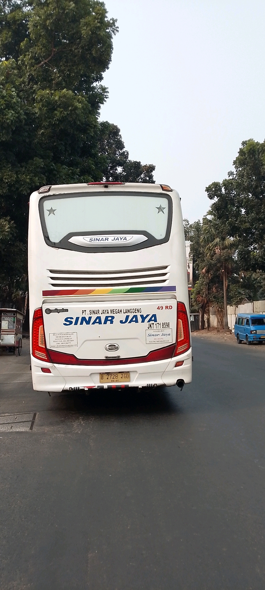 The image shows the rear view of a white bus with "SINAR JAYA" branding. The bus is parked on a street in Bekasi, Indonesia. The bus has a license plate that reads "B 7728 TGO". A blue minivan is visible in the background on the right side of the image. Trees line the street on both sides. A small food cart is parked on the left side of the image. The sky is overcast.