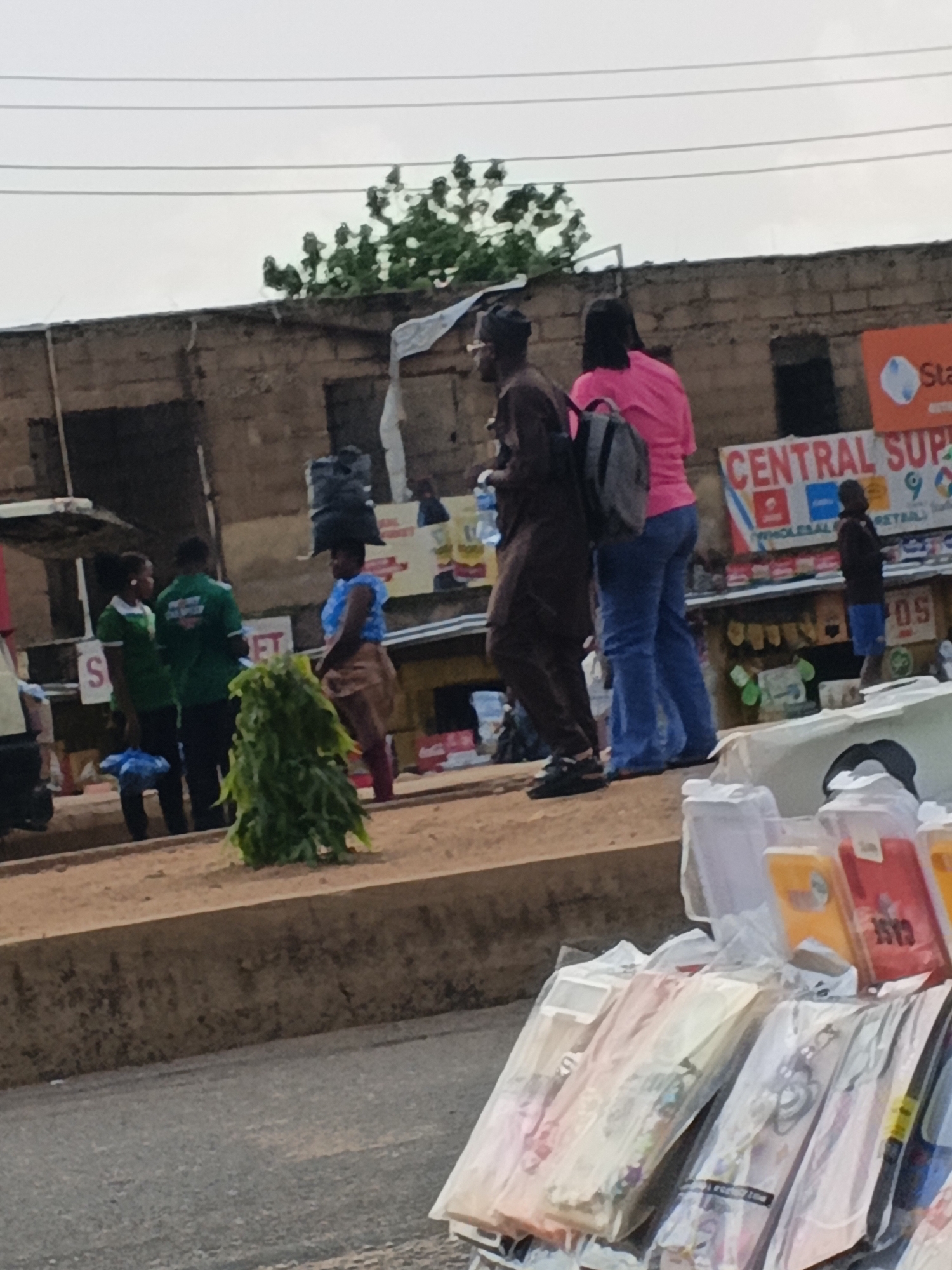 The image depicts a street scene in Bukuru, Nigeria, likely during the daytime under an overcast sky. The foreground features a display of mobile phone cases and accessories, packaged in clear plastic and arranged on a surface. The items are colorful and varied, suggesting a retail setting.
Behind this display, a group of people are visible in the middle ground. Several individuals are walking or standing near stalls that line a building. One person, a woman, is carrying a large black bag balanced on her head, a common sight in many African markets. She is wearing a blue top and a brown skirt.
Further back, a man in a dark brown tunic and trousers walks alongside a woman in a bright pink top and blue jeans, who is carrying a backpack. To their left, two individuals in green shirts are seen near a stall with a sign featuring the letter "S". The building behind them appears to be constructed of rough brick, with openings that suggest shops or market stalls. A large orange sign with white lettering is visible, partially obscured, with the words "CENTRAL SUP..." and "WHOLESALE & RETAIL" legible. Another sign with "POS" is also visible.
The overall scene suggests a busy marketplace or street with ongoing commercial activity. The presence of both vendors and customers, along with the variety of goods displayed, points to a typical day in a Nigerian urban or semi-urban setting. The weather appears to be cloudy, as indicated by the uniformly lit sky.