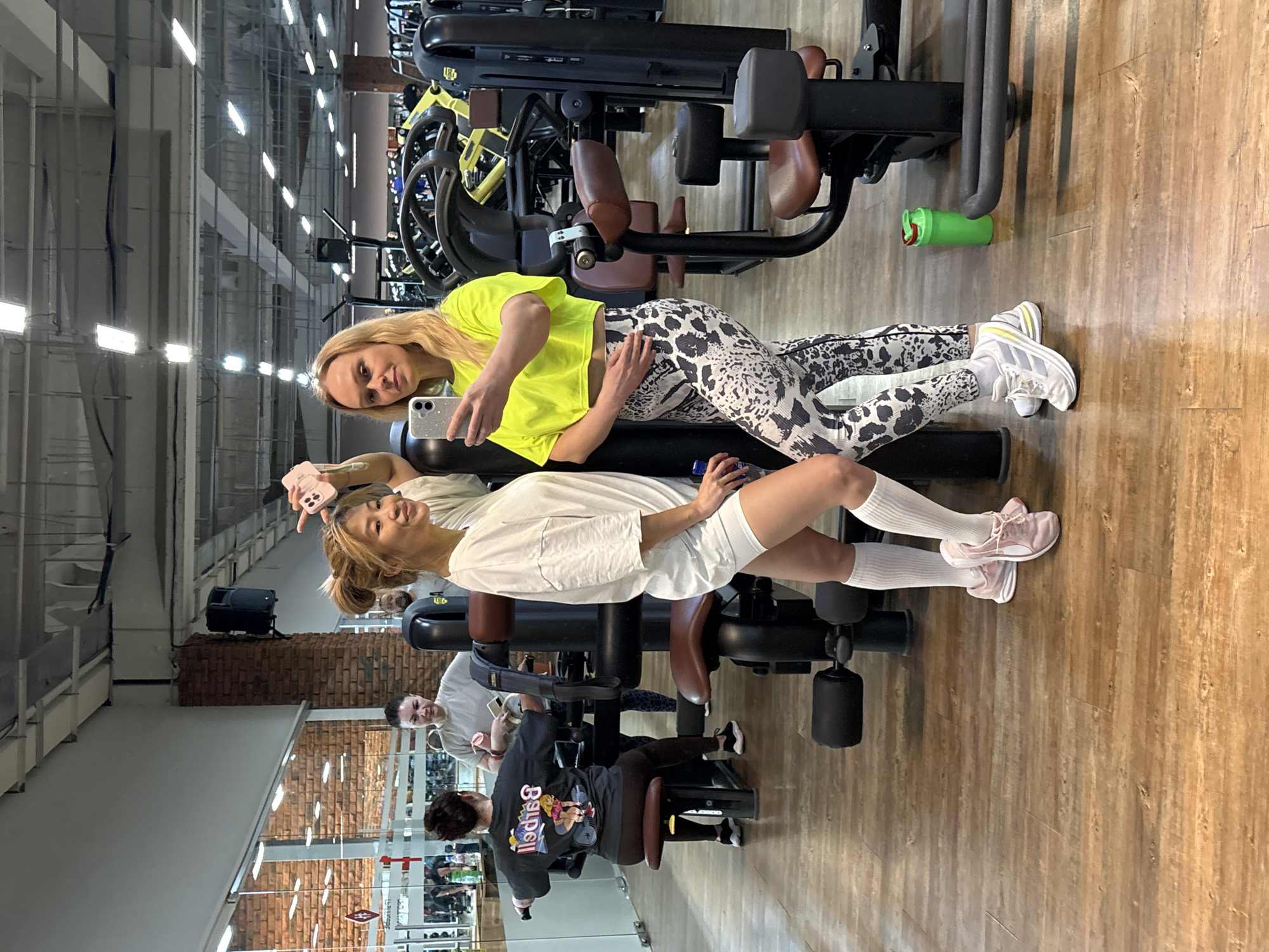 The image captures an indoor scene at what appears to be a fitness center or gym in Chaikovsky, Russia. Two women are the primary subjects, positioned on or near a piece of gym equipment. The woman in the foreground is wearing a white t-shirt, shorts, and white socks with pink sneakers, holding a pink phone. The other woman is wearing a neon yellow top and patterned leggings with white sneakers, holding a white phone. The two appear to be taking a selfie or photo together. Several other individuals are present in the background. One is using a machine. Other gym equipment is visible, along with architectural details like brick walls and large windows, suggesting a spacious, well-lit environment. A green bottle is next to the leg press machine.