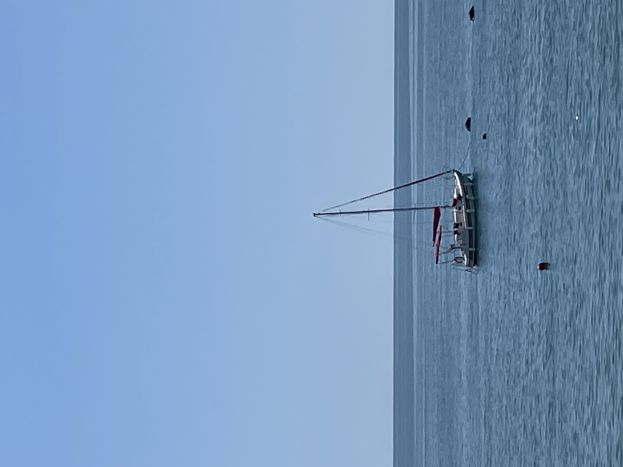 An open body of calm blue water stretches to the horizon under a clear, pale blue sky. In the middle distance, a white sailboat with red trim and a red canopy is anchored. The boat has a single mast and appears to be empty. Several dark buoys are visible in the water around the sailboat. The scene is likely the Black Sea coast near Sochi, Russia, given the calm water conditions and typical boating activity.