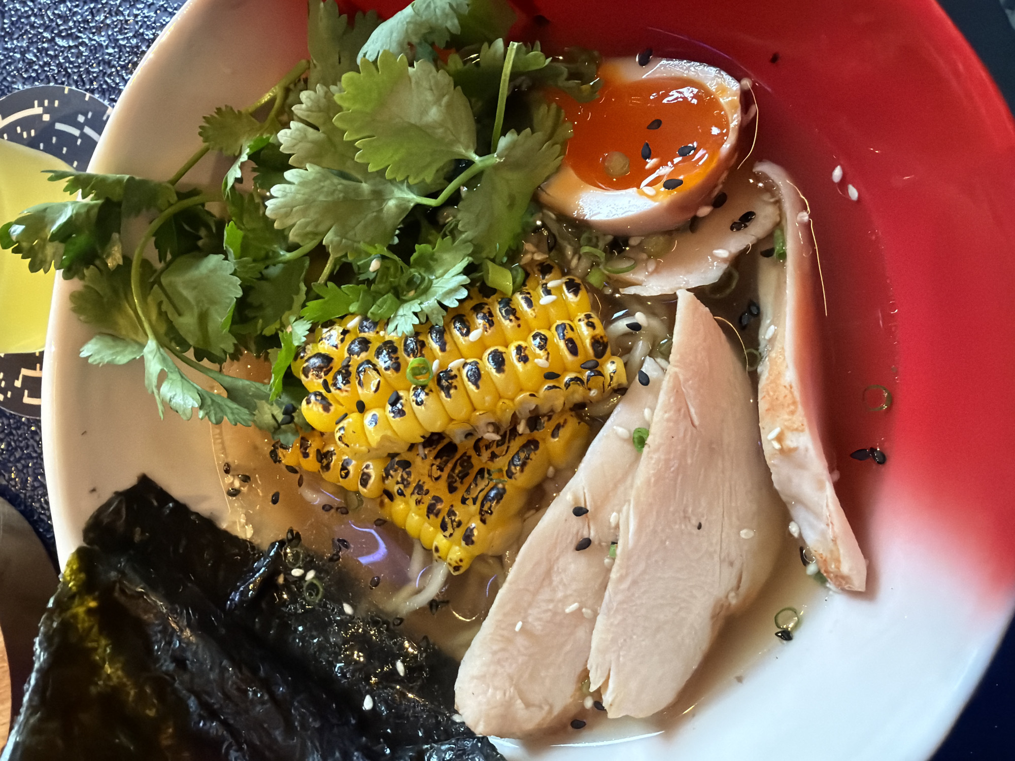 A close-up, overhead shot shows a bowl of chicken ramen. The ramen broth is clear with a golden hue. Sliced cooked chicken breast, grilled corn on the cob cut into three pieces, a halved soft-boiled egg with a bright orange yolk, and a sheet of dark green nori seaweed are the main toppings. The bowl has a white interior with a red exterior rim. Scattered black and white sesame seeds and chopped green onions are visible throughout the dish. The background is blurred, but appears to be a table with a dark blue textured surface and part of a yellow drink in a glass. There are no people or animals present in the image. The location is unknown.