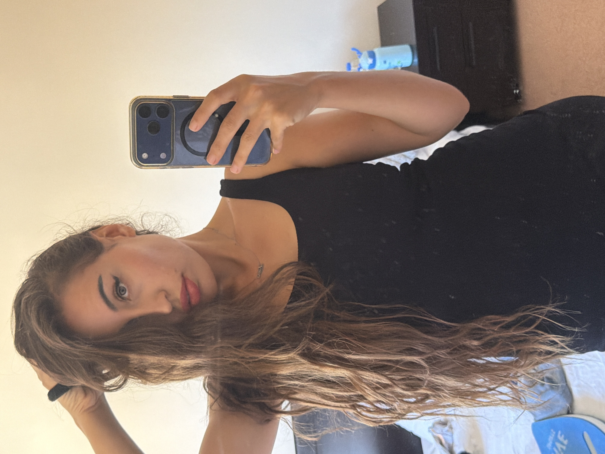 A young woman is taking a mirror selfie in a bedroom. She is wearing a black tank top and has long, wavy brown hair that is partially covering her face. Her left hand is raised, holding her hair. She is wearing light makeup, including winged eyeliner and pink lipstick. A silver necklace with a small pendant is visible around her neck. She is holding a dark blue smartphone with a gold-rimmed case in her right hand, pointed towards the mirror. The background shows a light-colored wall, a bed with white linens on the left side, and a dark dresser with a blue water bottle on top on the right side. The overall scene suggests a casual, personal moment captured in a domestic setting. The image does not contain specific visual cues to confirm the location as Tersefanou, Cyprus.