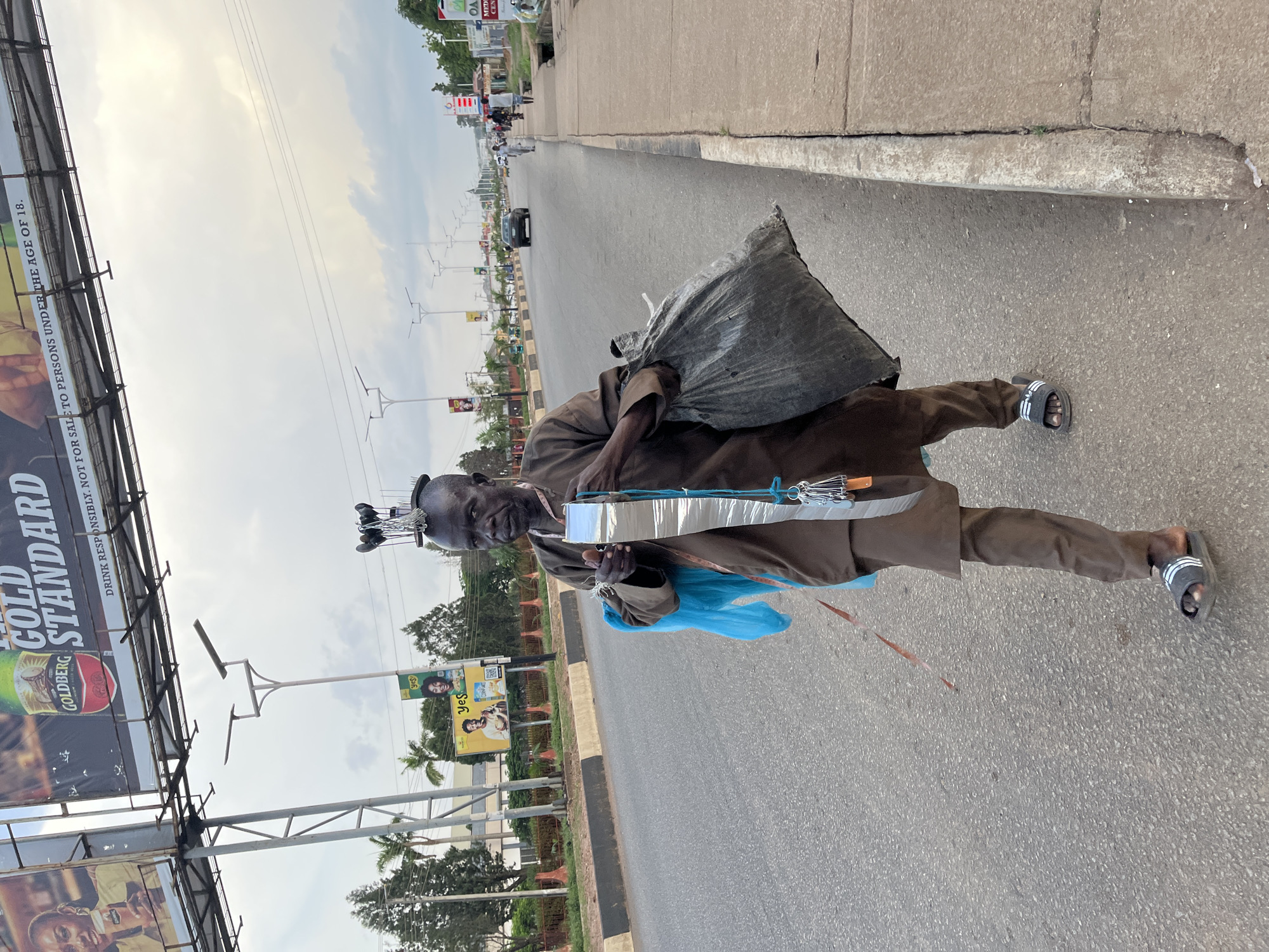 The image captures a street scene in Jos, Nigeria. In the foreground, a man walks along the side of a road. He is dressed in a brown traditional outfit and is carrying a large black trash bag in his right hand. He also holds a blue plastic bag and a long, reflective tape with keys attached to it in his left hand. His head is adorned with several items, including what appear to be metal objects and possibly a comb. He is wearing dark sandals with white stripes.
The road is paved and appears to be a busy thoroughfare with a concrete sidewalk on the right. Several cars are visible in the distance, indicating traffic. Various billboards and streetlights line the road, some displaying advertisements. The sky is overcast. The overall impression is a candid street photograph depicting everyday life in an urban environment.