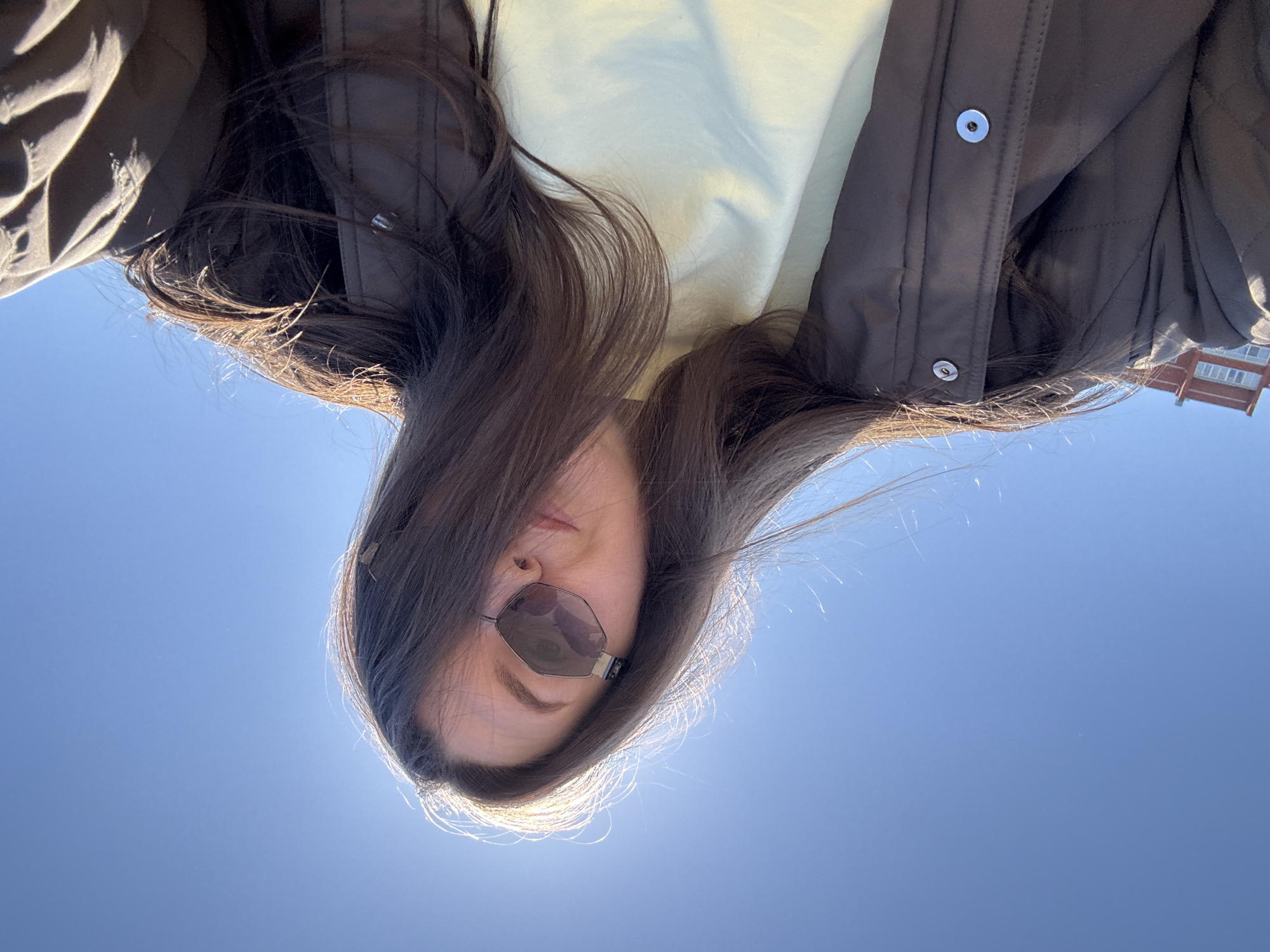 A woman with long brown hair and sunglasses is pictured from a low angle, looking down at the camera. She is wearing a light yellow shirt under a brown quilted jacket with silver buttons. The background is a clear blue sky. A building is visible in the upper right corner of the image, consistent with urban architecture of the city of Chaikovsky, Russia.