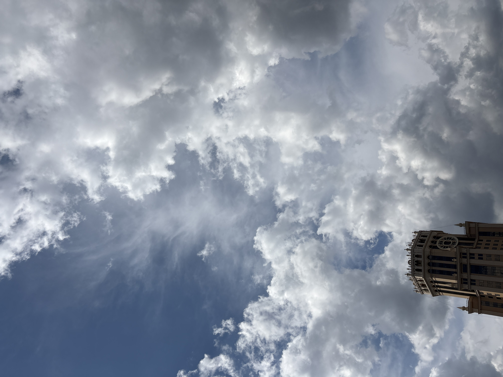 The image captures a low-angle view of the iconic Moscow State University main building, a Stalinist skyscraper, set against a dramatic sky filled with an array of clouds. The building, with its distinctive tiered architecture and ornate crown, is partially obscured by the dynamic cloud formations. The sky displays a gradient from deep blue to lighter shades, with wispy cirrus clouds mingling with heavier, darker cumulus clouds, suggesting an approaching weather change. The overall atmosphere is one of imposing architecture meeting the grandeur of nature.