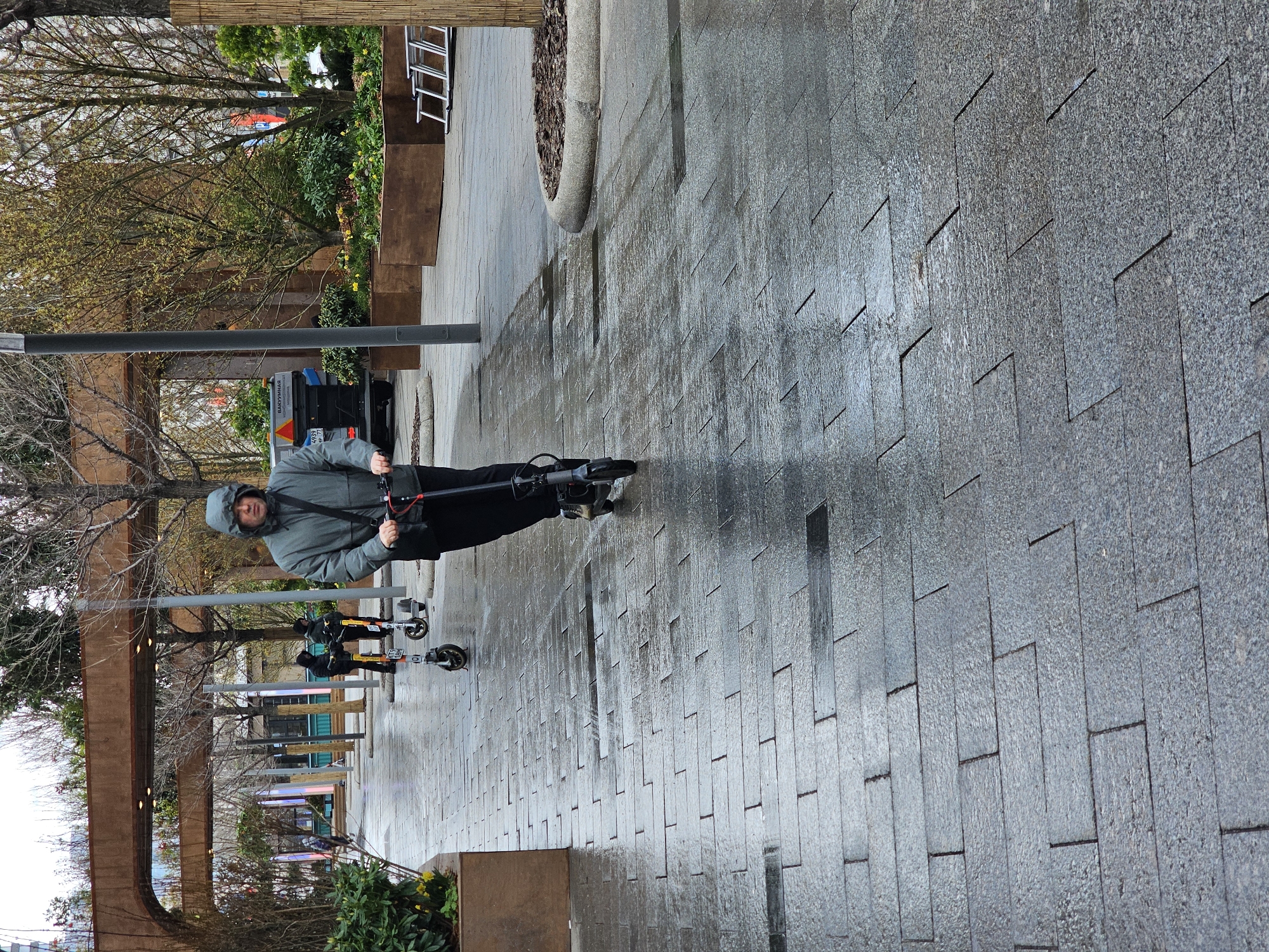 A medium shot captures a man riding an electric scooter on a paved walkway. The man is dressed in a gray hooded jacket and dark trousers. He is traveling to the left.
The walkway is made of gray rectangular stones arranged in a herringbone pattern, and appears wet, reflecting the ambient light. Along the left edge of the walkway, there are lampposts and trees, creating a path or walkway. Other parked electric scooters can be seen further down the walkway.
In the background, there is a small truck.
Based on the architectural style of the walkway and lampposts, this image appears to be captured in a modern public space, possibly a park or pedestrian area. The location is potentially in the city of Moscow, Russia.