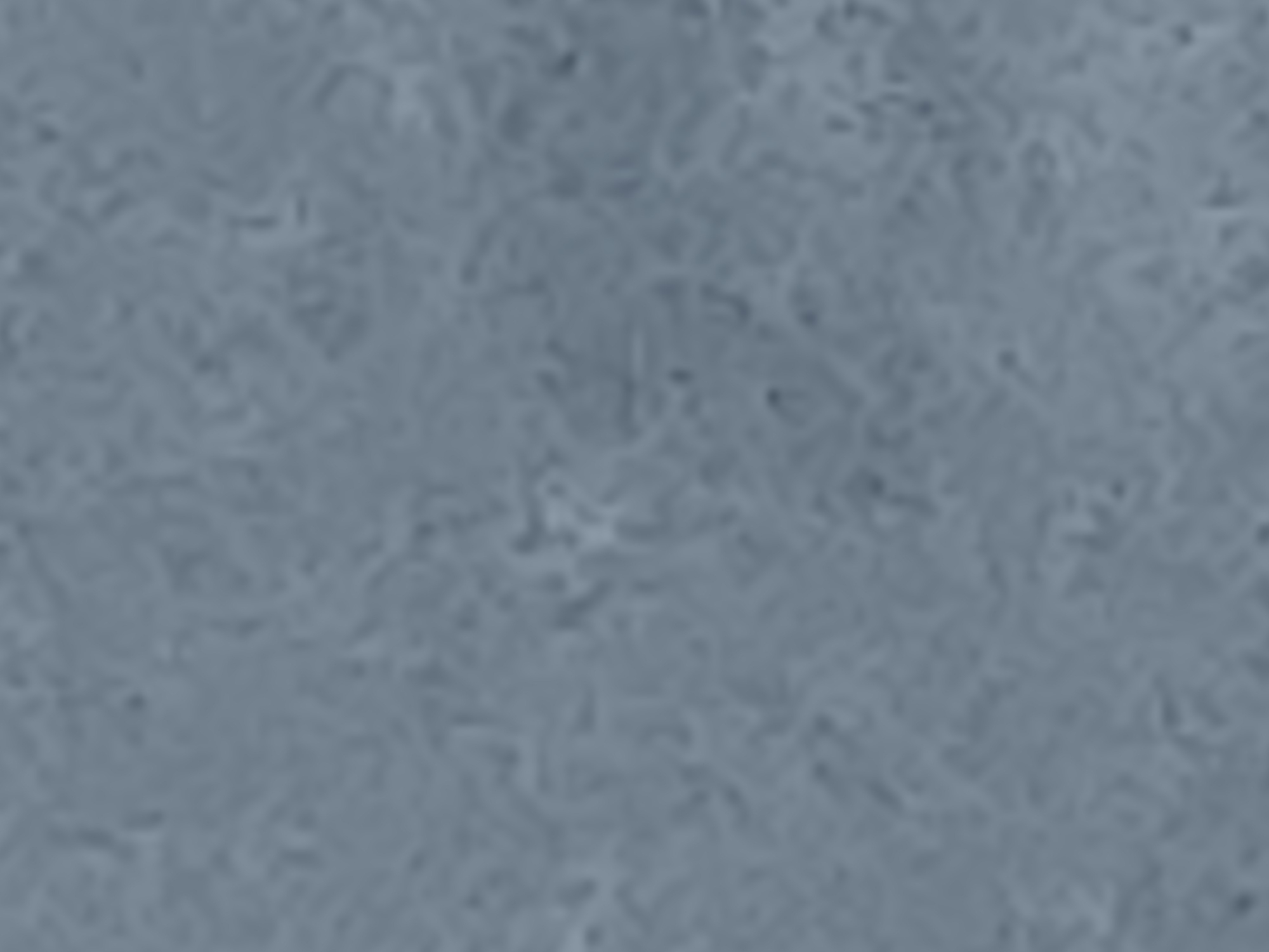 The image is a close-up, out-of-focus shot of a textured surface, appearing to be a pale gray or blue-gray material with subtle variations in tone and pattern. The texture suggests it could be fabric, a painted surface, or natural material like stone or wood, but it is too indistinct to determine with certainty. There are no discernible objects, people, or environmental features visible in the image. The lighting appears diffuse, with no harsh shadows.
