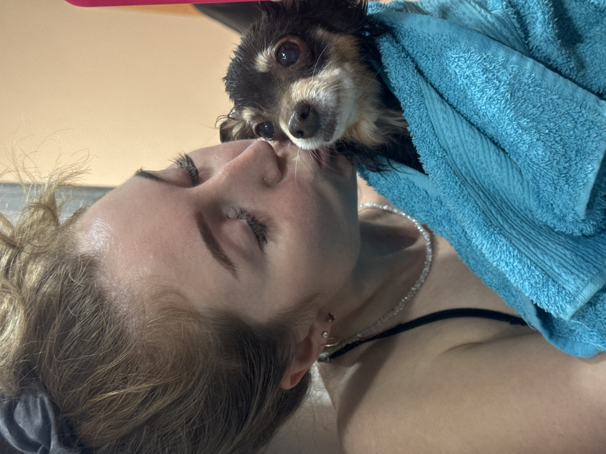 A woman lies on her back while a small, wet dog wrapped in a blue towel licks her face. The woman has fair skin, closed eyes, and light brown hair pulled back. She wears a thin black strap on her shoulder and a pearl necklace. The dog is mostly black with brown and white markings. The background is a peach-colored wall with a pink object out of focus at the top. The location cannot be determined from the image.