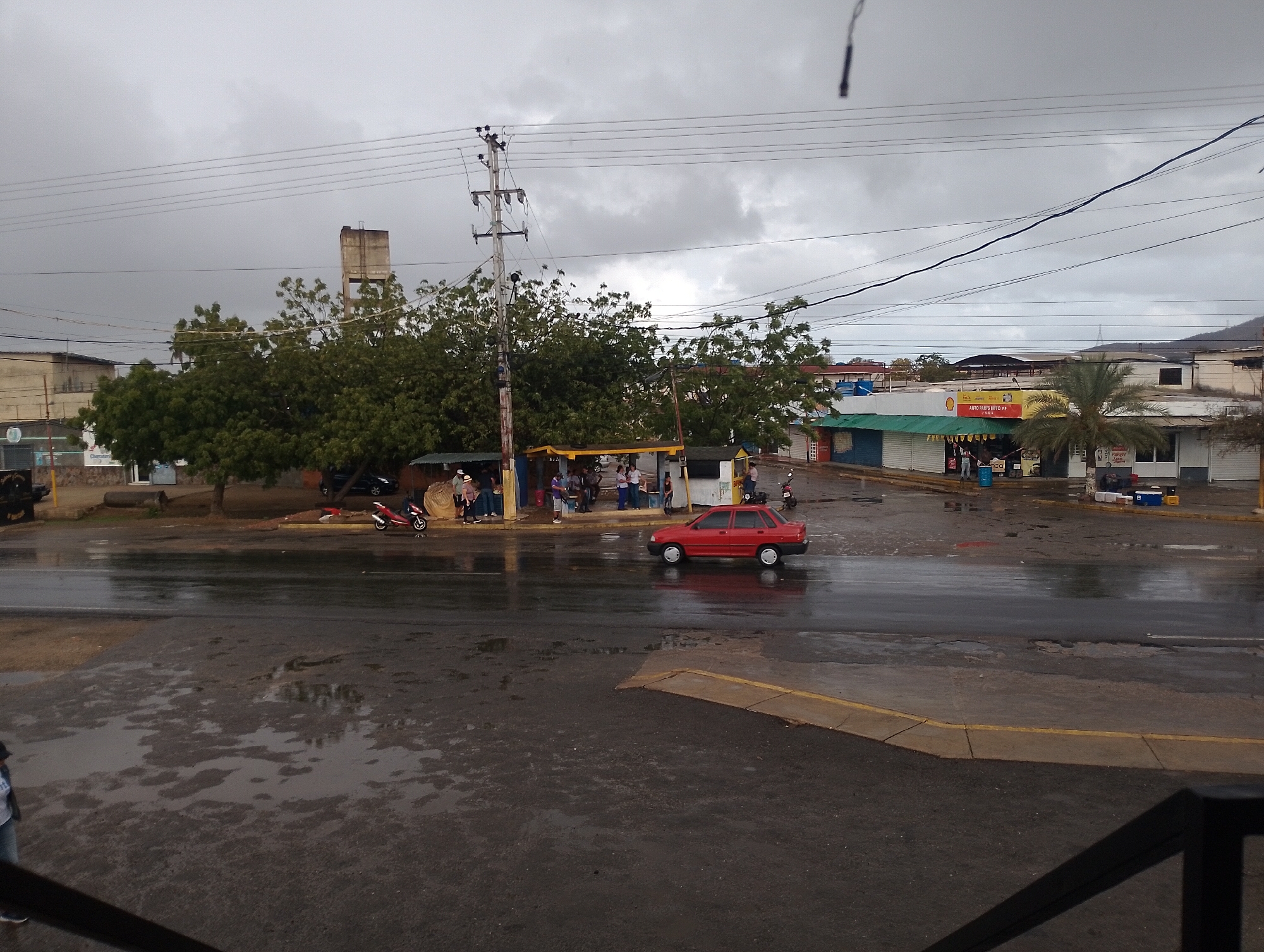 This image depicts a street scene in El Espinal, Venezuela, under an overcast sky, with the ground visibly wet from recent rain, displaying numerous puddles. In the background, a multi-story concrete tower structure rises above a dense canopy of green trees. A complex network of overhead power lines traverses the upper portion of the frame, anchored by a prominent utility pole on the left. The midground features various low-rise commercial buildings lining a street. One notable building on the right has a green awning, a yellow "SHELL" sign, and another sign identifying "AUTO PARTS AUTO K.P." A palm tree stands in front of this building. Several individuals are visible congregating on the wet pavement and sidewalks in front of what appear to be makeshift market stalls or small shops. A red two-door sedan is centrally positioned on the wet asphalt road, while at least two motorcycles are parked near the groups of people on the sidewalk. The foreground consists of dark, wet asphalt, reflecting the grey sky, with a yellow curb marking the edge of a paved area on the lower right. The lower legs of a person are visible in the bottom-left corner.