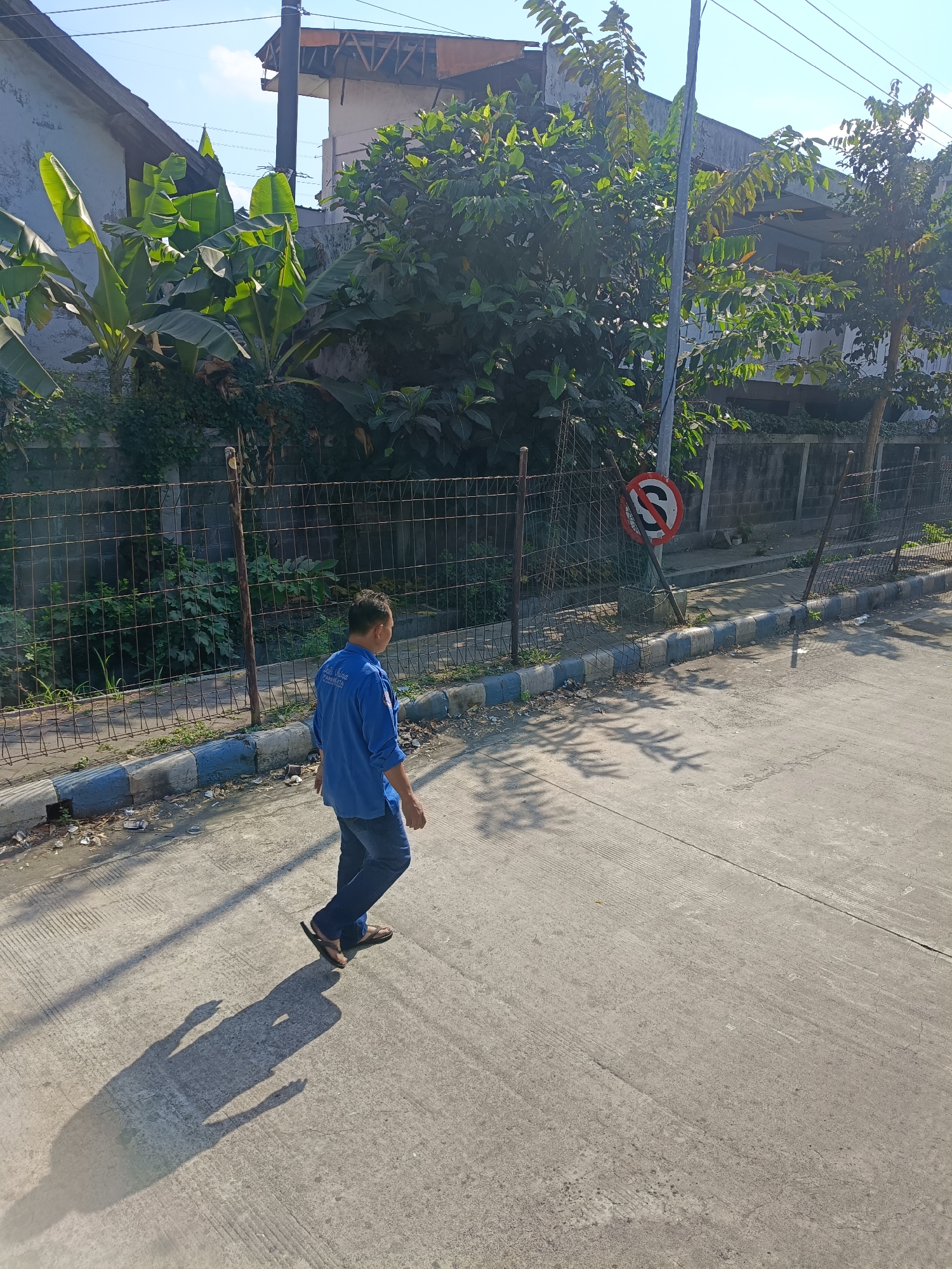 A man in a blue long-sleeved shirt and jeans walks on a concrete road in Kedungrejo, Indonesia, casting a long shadow. He is wearing flip-flops and appears to be walking away from the camera. A wire fence runs along the left side of the road, behind which are overgrown plants and some residential buildings with tiled roofs. A traffic sign, partially obscured by a pole, is visible on the fence, indicating a prohibition. The sky is clear and blue, suggesting a sunny day. The environment appears to be an urban or semi-urban setting.