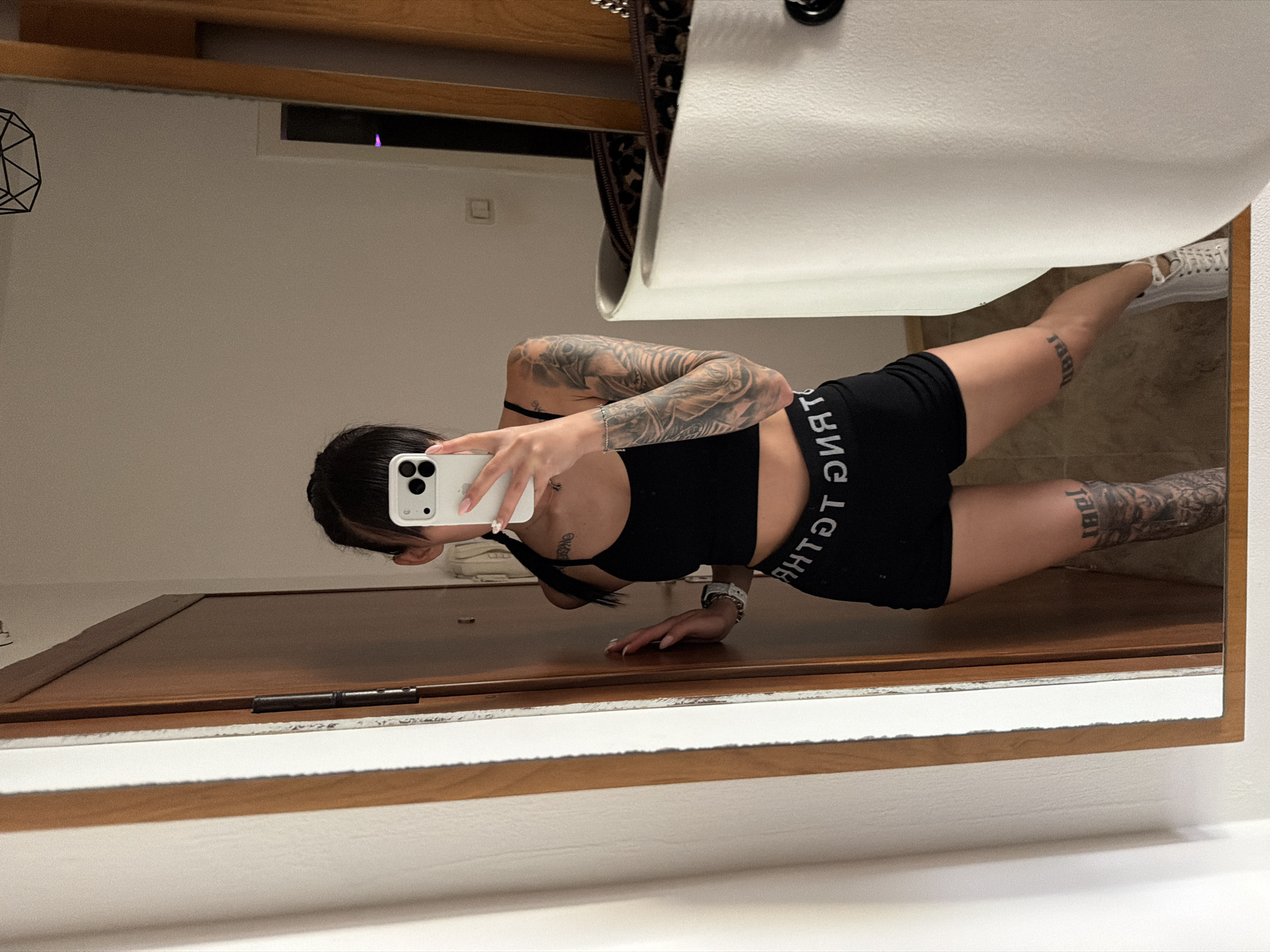 The image shows a young woman with dark hair and bangs taking a mirror selfie indoors. She is wearing a black crop top and black shorts with white text. She has tattoos on her arms and legs. In the background, a wooden door is visible, and a mirror reflects a portion of the room. A white laundry basket or hamper is also partially visible in the foreground. The overall setting suggests a residential environment, potentially an apartment or house. There is no specific indication of the city of Plovdiv, Bulgaria, or any landmarks that would place the location there.