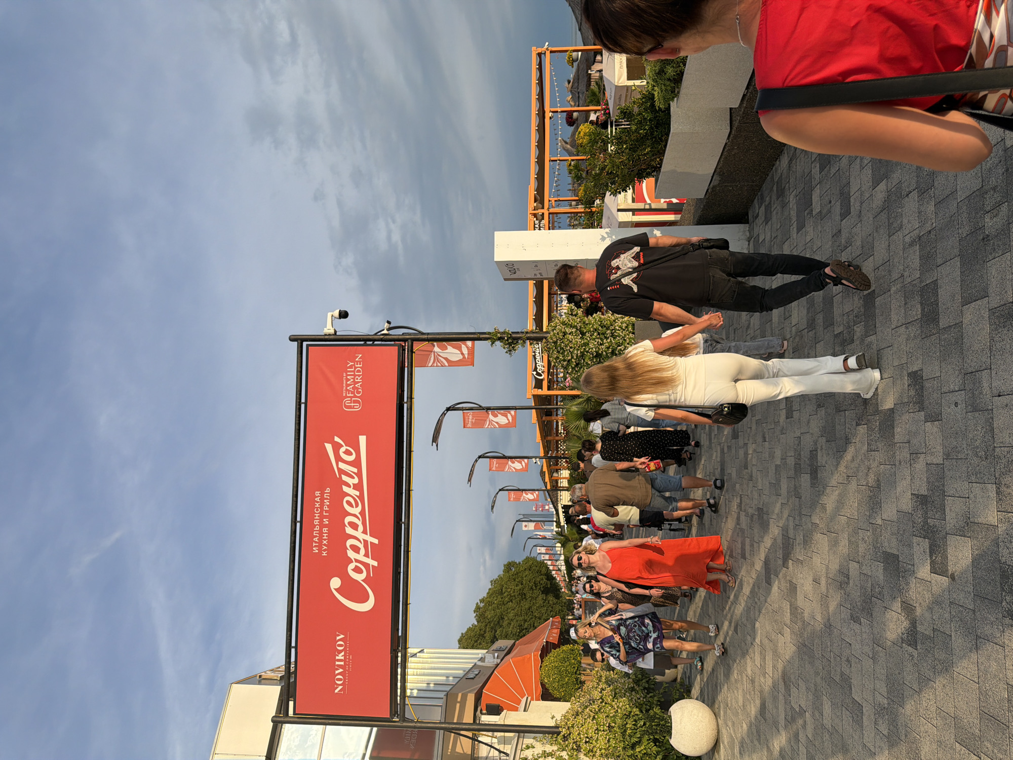 In Sochi, Russia, a pedestrian walkway is filled with people strolling under a cloudy sky. Overhead, a large red banner displays "NOVÍKOV" and "Sorrento" in prominent white lettering, indicating a restaurant or establishment. The walkway is paved with gray rectangular tiles, and shadows from the setting sun stretch across the ground. To the left, a building with orange awnings and lush green bushes lines the path. To the right, modern outdoor seating areas with white structures and wooden roofs are visible. The people, dressed in casual summer attire, walk in various directions, some in pairs, others alone or in groups. A woman in a bright orange dress walks towards the viewer, while a couple holding hands walks away. The scene captures a lively evening atmosphere in a resort area.