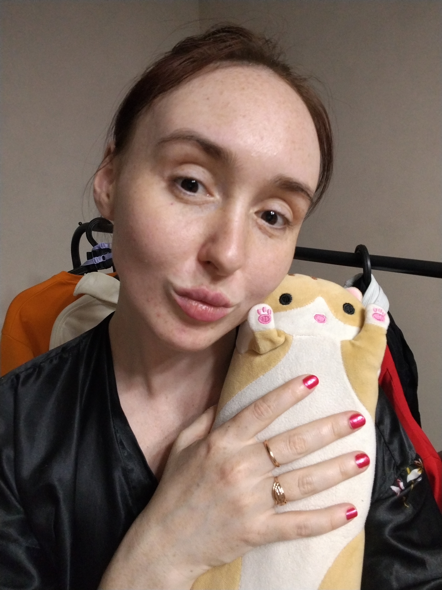A woman with light skin, dark hair pulled back, and visible freckles on her forehead is captured in a close-up, chest-up selfie. She has dark eyes and is making a playful expression with puckered lips. She wears a black, possibly satin, top or robe. Her fingernails are painted bright red, and she has two gold-colored rings on her left hand: one on her ring finger and another, more intricately designed, on her middle finger.
She is holding a light brown and white plush toy that resembles a hamster or small rodent. The toy has black eyes, a pink nose, and pink paw prints on its upright paws.
The setting appears to be indoors, against a light-colored wall. Behind her, a black clothing rack is visible, from which an orange garment, a white garment, and some darker clothes are hanging. The overall impression is informal and candid. The provided location context is Izhevsk, Russia. There is no visible text in the image.