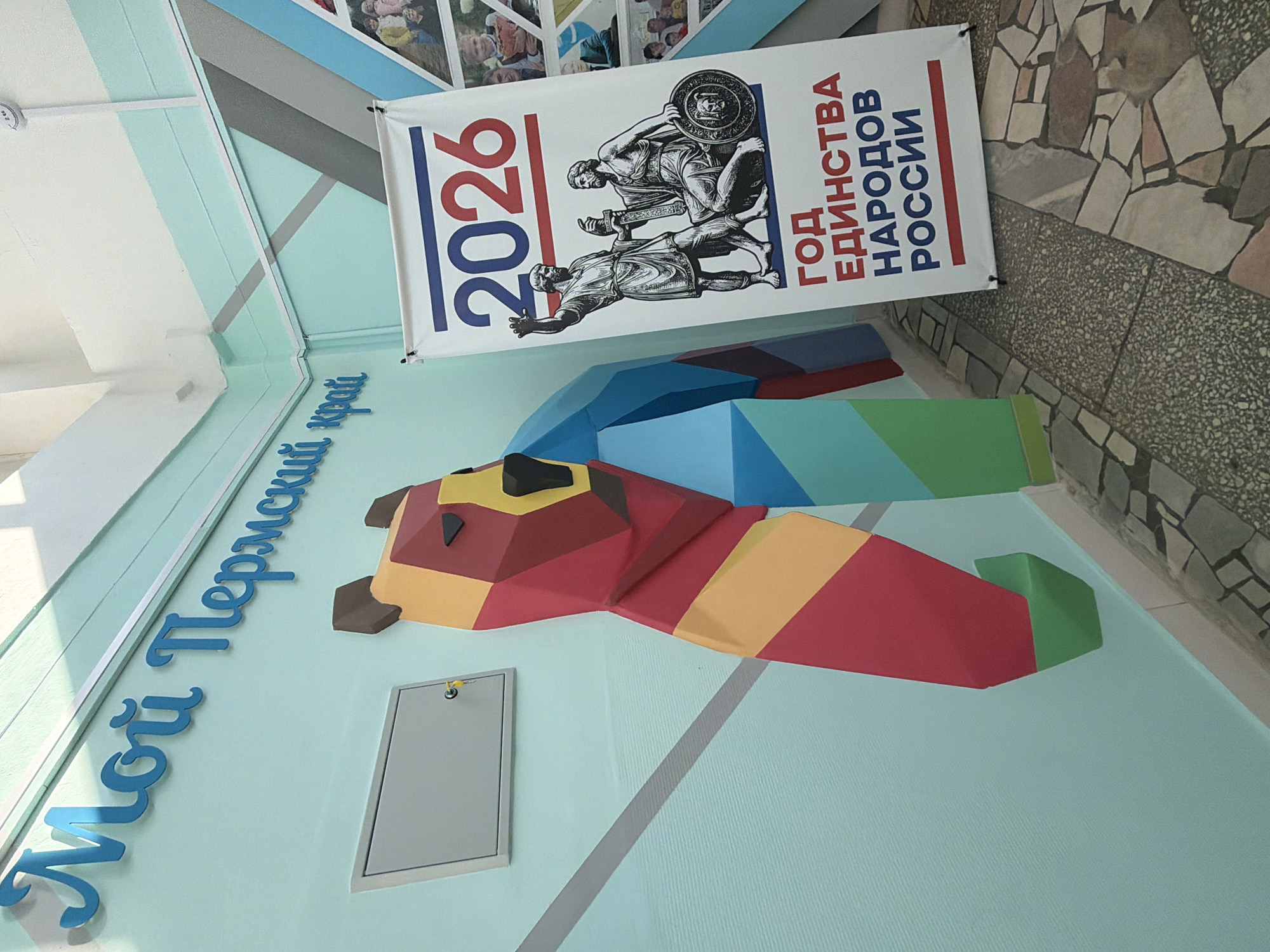 A vertically-oriented medium shot captures an indoor wall display. On the wall is text in Cyrillic script reading "Мой Пермский край" in a sky blue hue. Above the text is a 3D polygonal artwork of a bear in red, yellow, brown, and black. This artwork is accompanied by a geometrically-shaped piece in blue and green, suggesting an abstract representation. To the right of the bear's head is a grey metal access door. Above the bear is a white banner proclaiming "2026 ГОД ЕДИНСТВА НАРОДОВ РОССИИ." The banner also features a sketch of two wrestlers in a Greco-Roman style. Overhead, a ceiling with white and blue panels is visible, and along the right side, the display is adjacent to an area with stone flooring. This scene is located in the city of Chaikovsky, Russia.