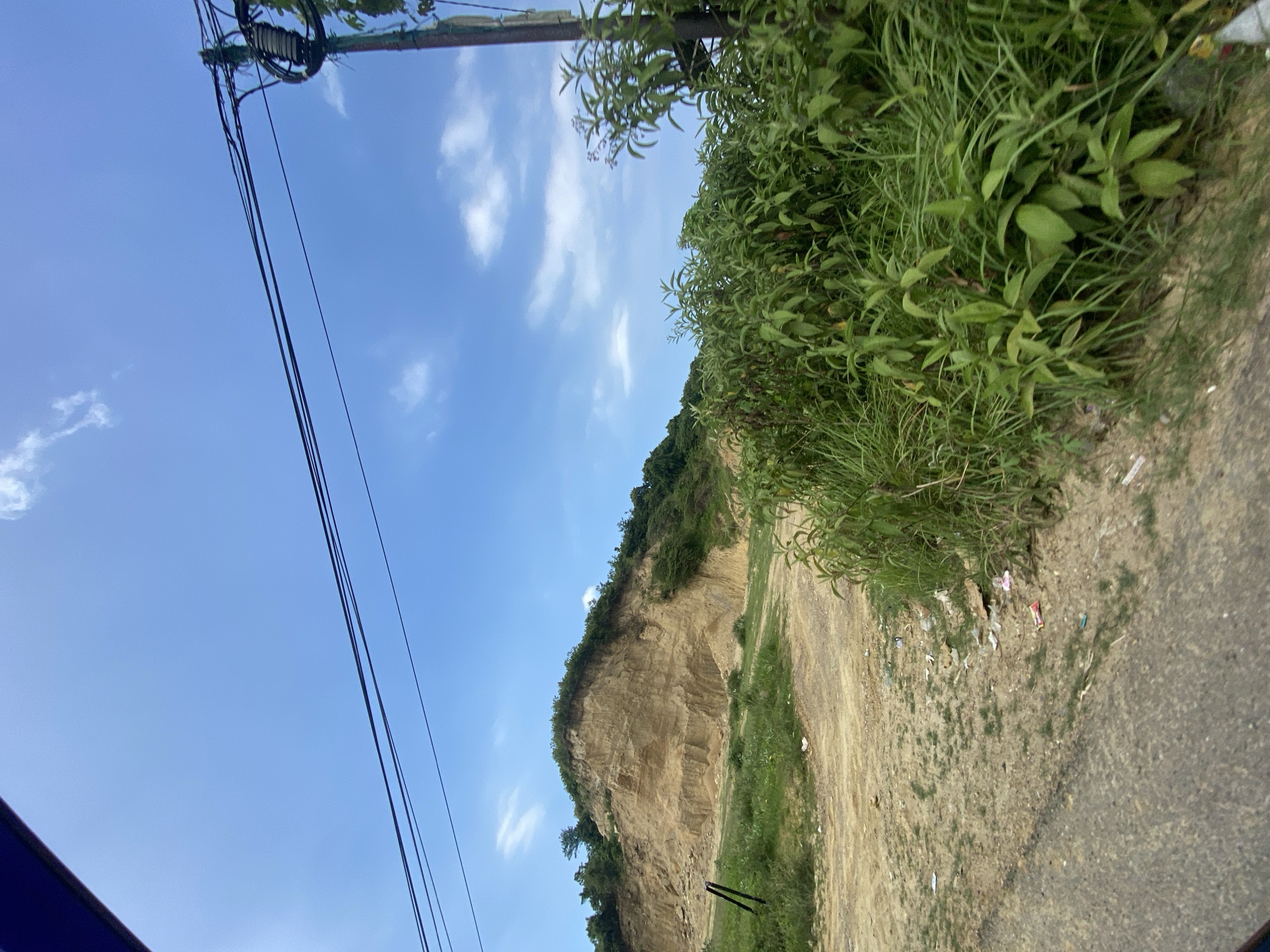 The image depicts a utility pole and power lines on the left, extending diagonally upwards. To the right, there's a cliff face or embankment partially covered with vegetation, including grass and shrubs. The ground appears to be a mix of dirt, gravel, and some trash. The sky is blue with scattered clouds. The location is Lhokseumawe, Indonesia, based on the prompt. There are no people visible in the image.