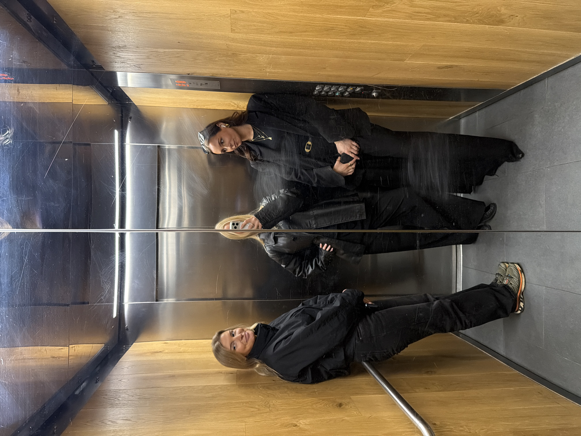 The image shows a reflection of two women taking a photo in an elevator. The elevator has wooden paneling on the top and bottom and stainless steel walls with a horizontal beam separating the upper and lower sections. The floor is gray. One woman is wearing a black blazer and sunglasses, and the other is wearing a black jacket. The location is likely within a building in London, United Kingdom, given the style of architecture and design.