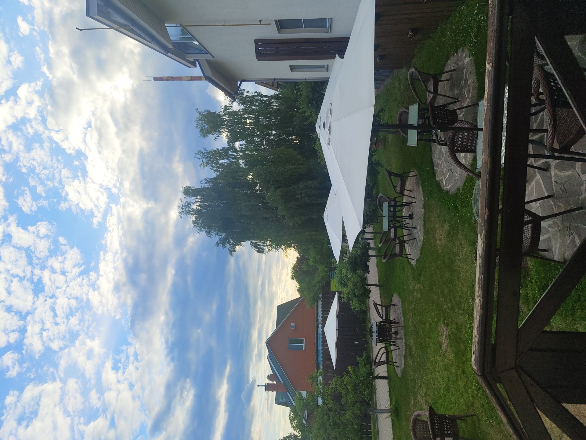 The image depicts an outdoor seating area with tables and chairs under large white umbrellas in a grassy yard. A brick house with a green roof is visible in the background, along with several trees and a wooden fence. The sky is cloudy with patches of blue. The scene appears to be a quiet, possibly residential or commercial establishment, in the city of Tsibino, Russia. There are no people or animals visible in the frame. The focus is on the architecture and the outdoor amenities.