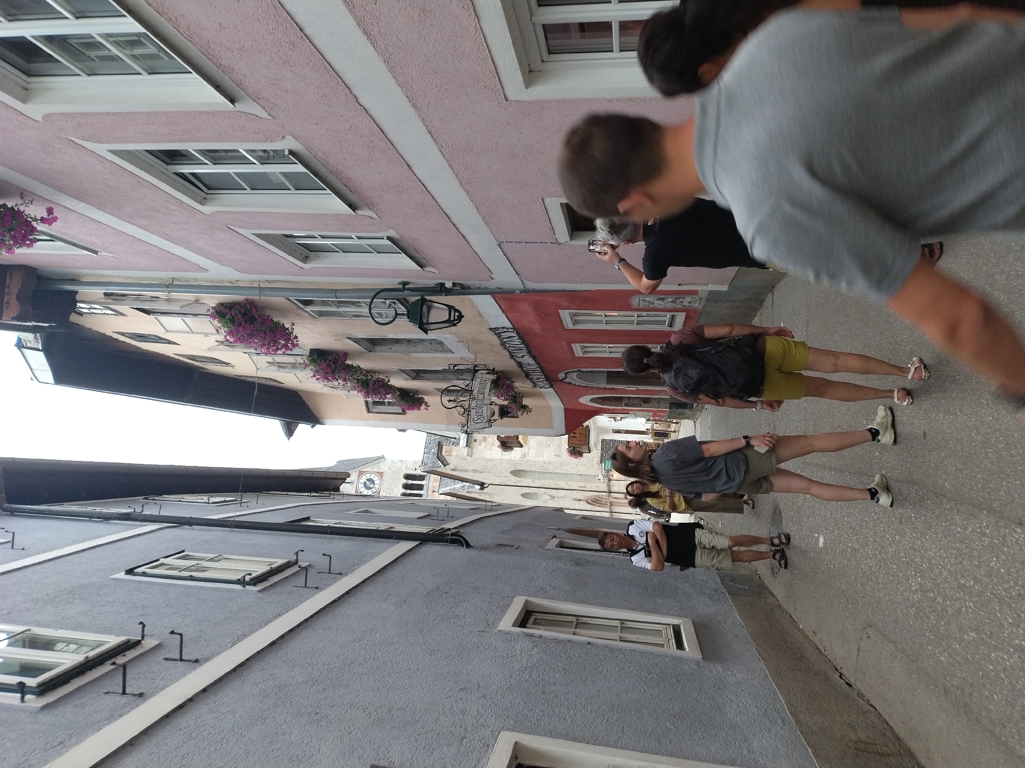 The image depicts a narrow cobblestone street in Hallstatt, Austria, lined with colorful, historic buildings. The architecture features pastel-colored facades, white-trimmed windows, and hanging flower baskets. On the left, a grey building has several windows with white trim. On the right, buildings display pink and cream colors. In the background, a church tower with a clock face is visible, suggesting a central location. Several tourists are present, walking along the street. One man in the foreground is wearing a grey polo shirt and shorts. Further down the street, a woman in a grey t-shirt and shorts walks with a backpack. Another man, wearing a black shirt and khaki shorts, stands to the side, looking towards the church. A woman in a yellow shirt and brown pants is also visible. The scene captures the charm of Hallstatt's pedestrian-friendly streets and its well-preserved historical architecture.
