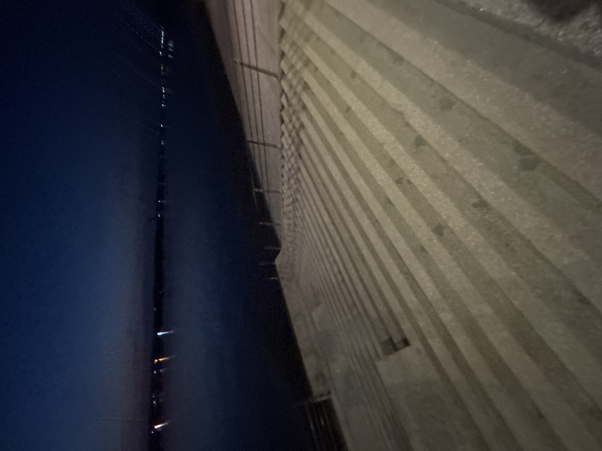 The image presents a nighttime view from the Kama Bridge in Perm, Russia. To the left, the dark expanse of the Kama River is visible. On the right, the concrete structure of the bridge is apparent, featuring a repeating pattern of horizontal lines, possibly sections of the bridge's supports or design elements. The image is angled, lending a dynamic perspective to the scene. Lights from the city reflect in the dark waters of the river.