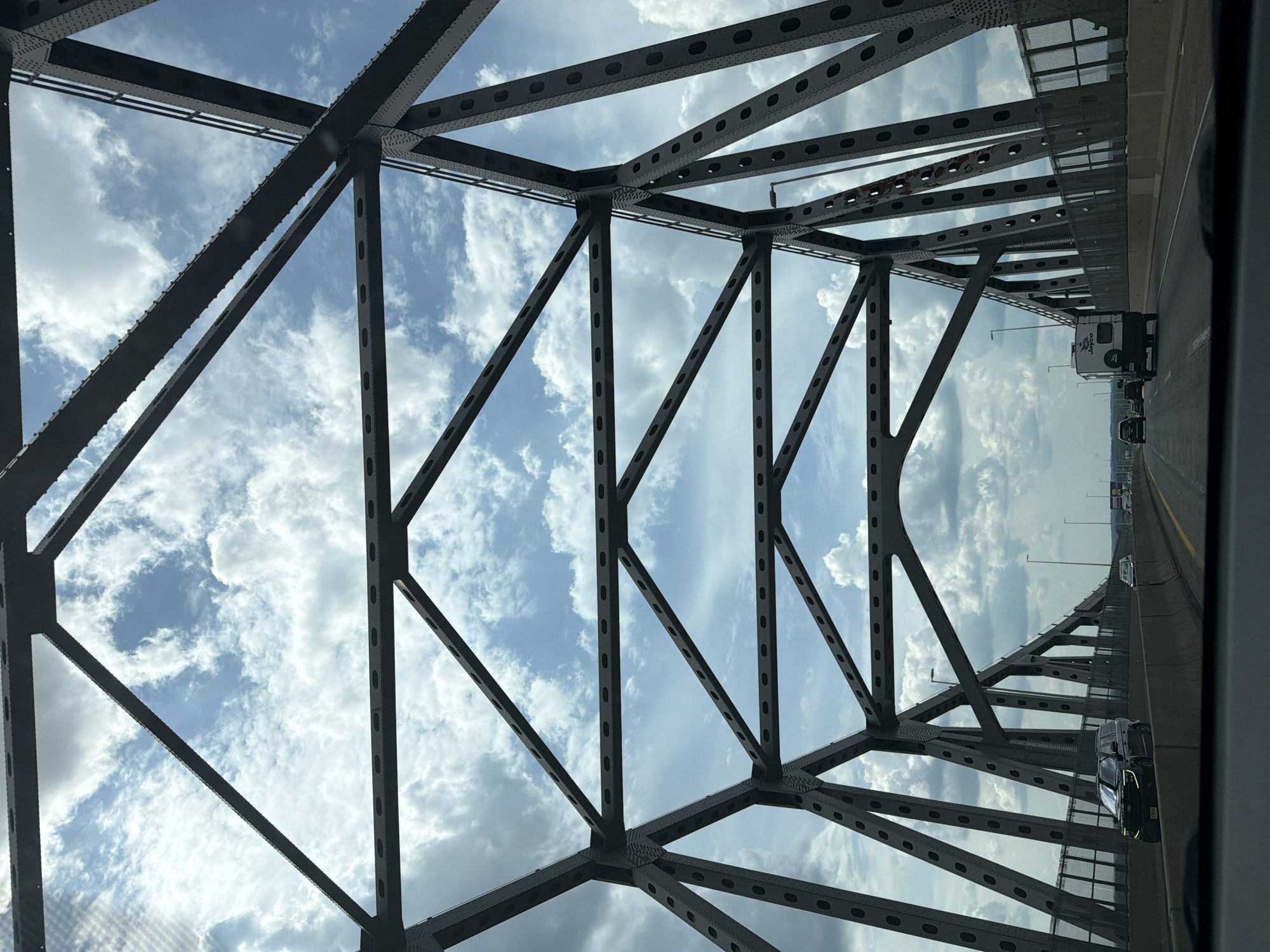 The image captures a view from a vehicle traveling across a large steel truss bridge, likely part of a highway in Bristol Township, United States of America. The perspective is looking forward and upward, with the intricate geometric patterns of the bridge's steel structure dominating the foreground and midground. Through the gaps in the metalwork, a bright blue sky filled with scattered white clouds is visible.

On the bridge deck, traffic is moving in the same direction as the viewer. Several vehicles are present, including a black SUV in the left lane with distinctive headlights, a white RV in the right lane, and other cars further ahead. Concrete barriers separate the lanes of traffic and the edges of the bridge. Streetlights are visible along the side of the bridge. The overall scene suggests a daytime journey on a well-trafficked roadway, framed by the imposing architecture of the bridge.